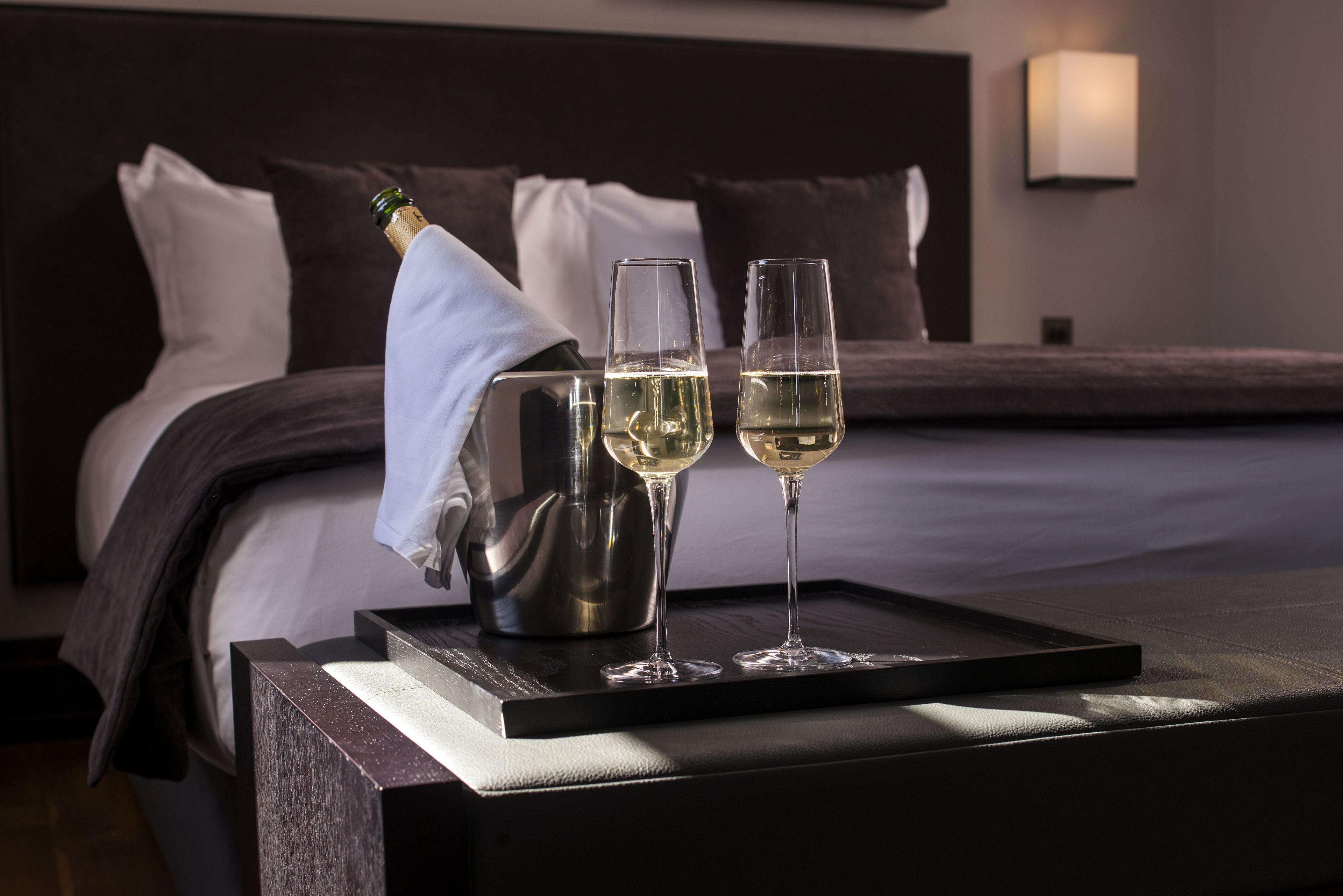 A neatly made bed with a dark headboard and pillows, a bedside table with a bottle and two glasses of white wine, and a wall-mounted lamp.