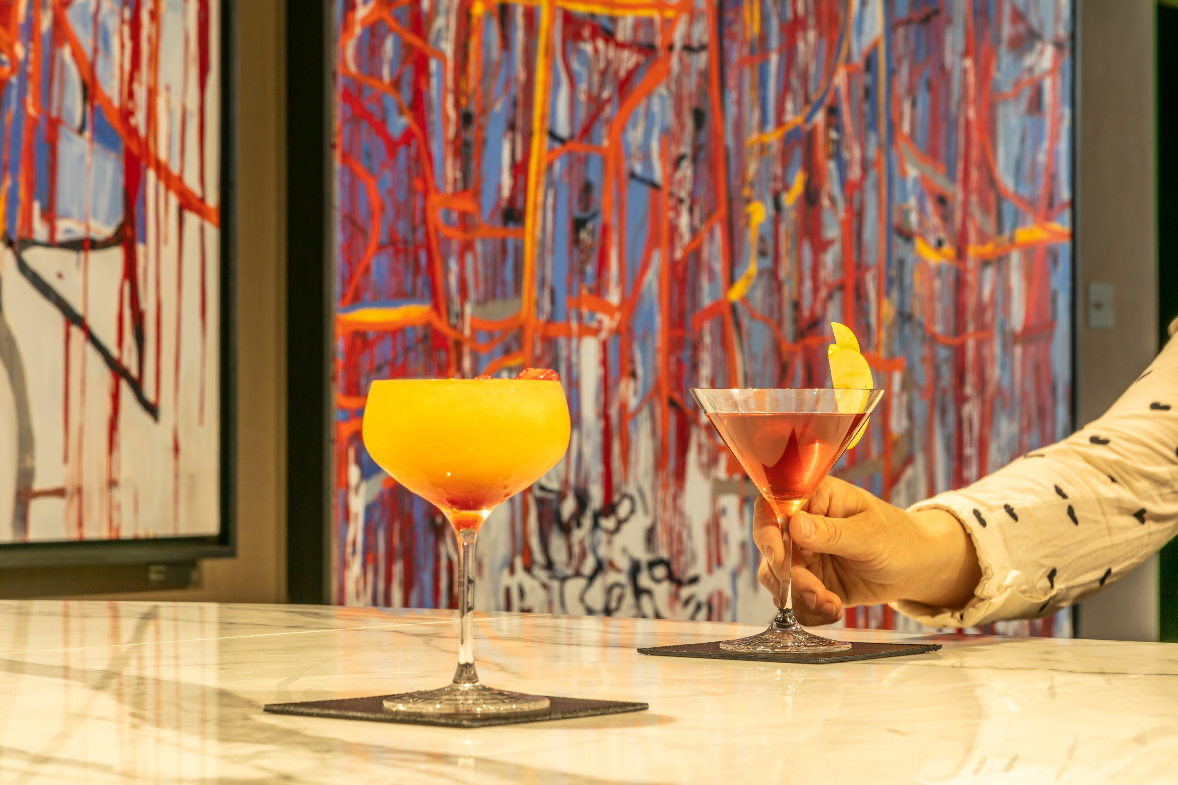 Two cocktails on a marble bar counter with a hand holding one, and abstract art in the background.