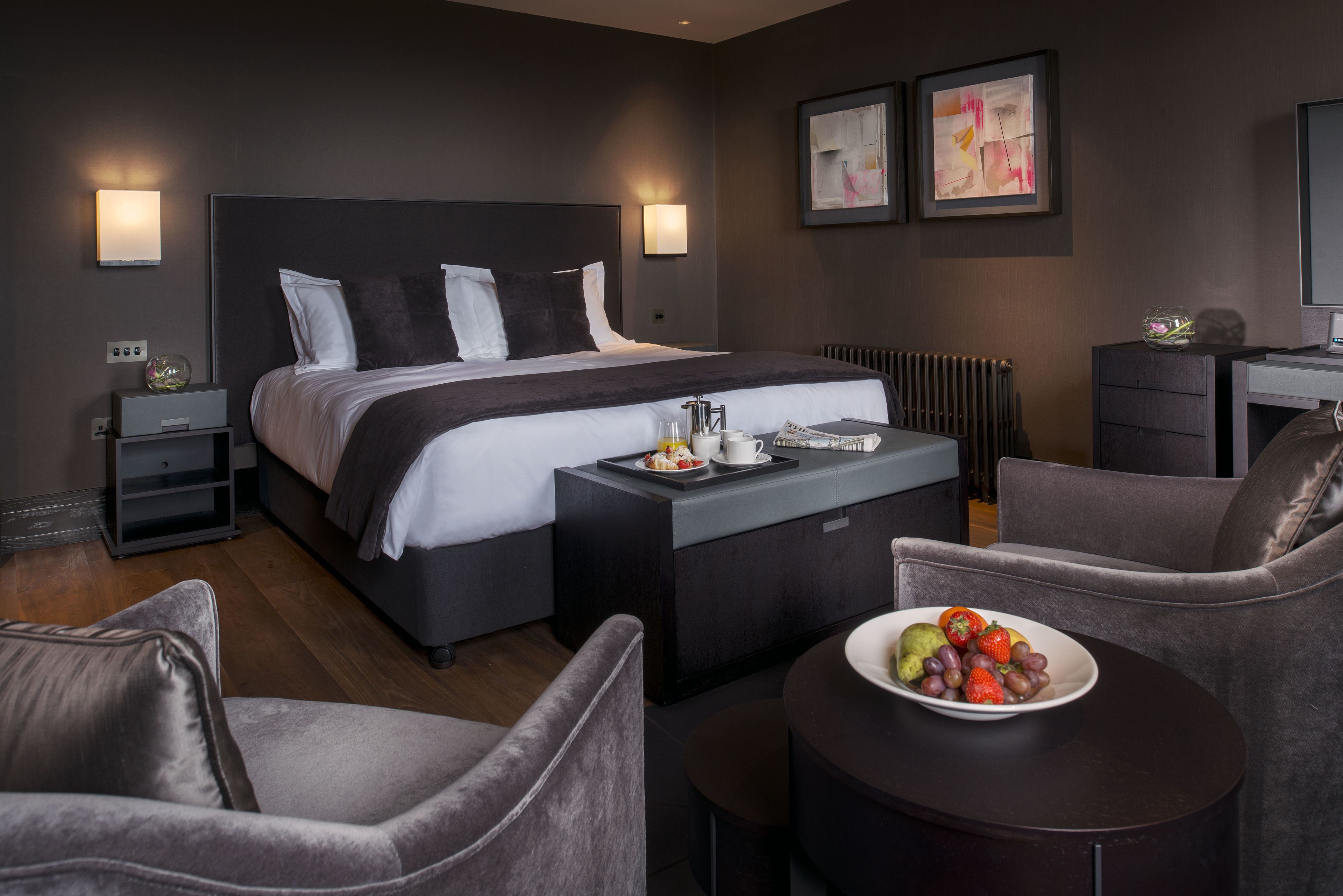 A modern hotel room with a large bed, bedside tables, a small desk, and a round table with fruit.