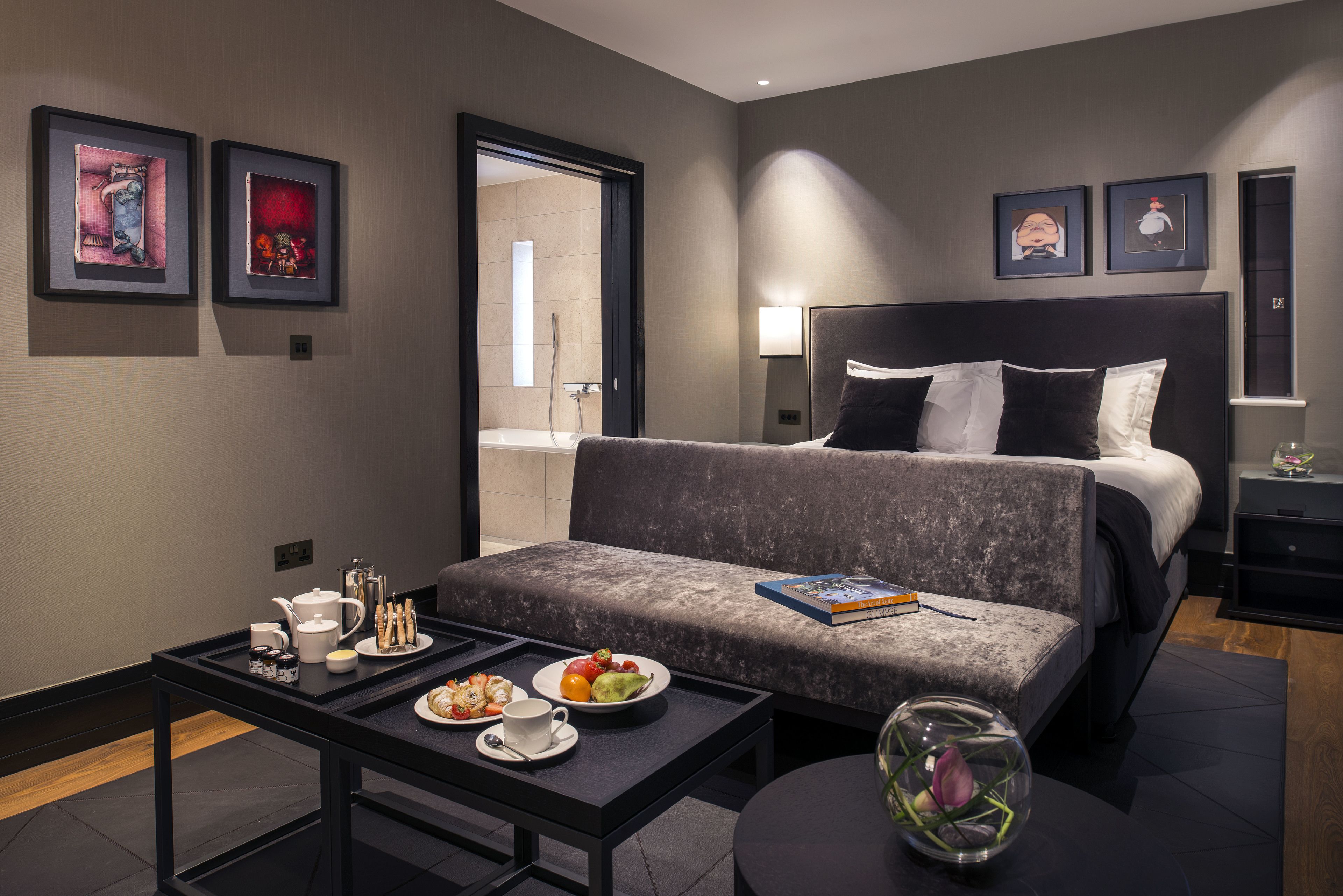 Ein modernes Hotelzimmer mit grauem Sofa, einem Bett mit schwarz-weißer Bettwäsche, einem Nachttisch mit Buch und einer Glasvase mit Blumen sowie einem Tablett mit Frühstücksgebäck.