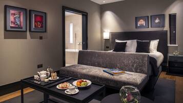 Ein modernes Hotelzimmer mit grauem Sofa, einem Bett mit schwarz-weißer Bettwäsche, einem Nachttisch mit Buch und einer Glasvase mit Blumen sowie einem Tablett mit Frühstücksgebäck.