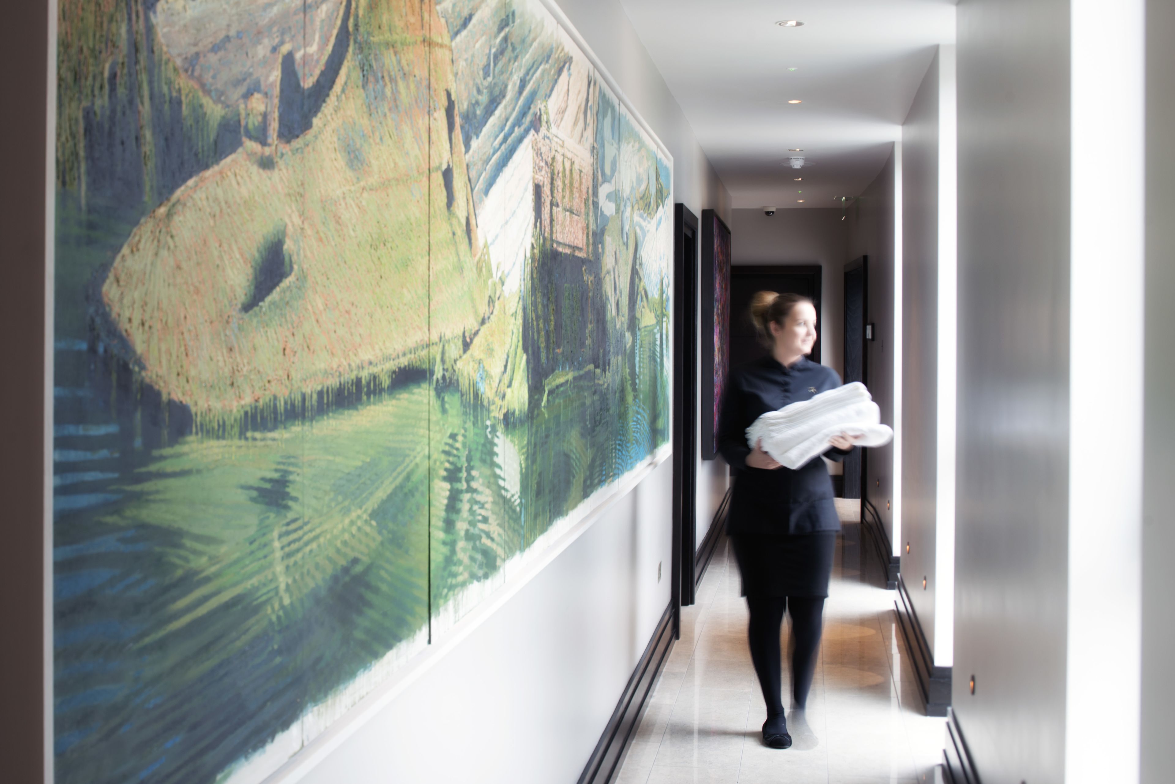 A hallway with a large abstract painting on the left wall, a person carrying a white object, and a door at the end.