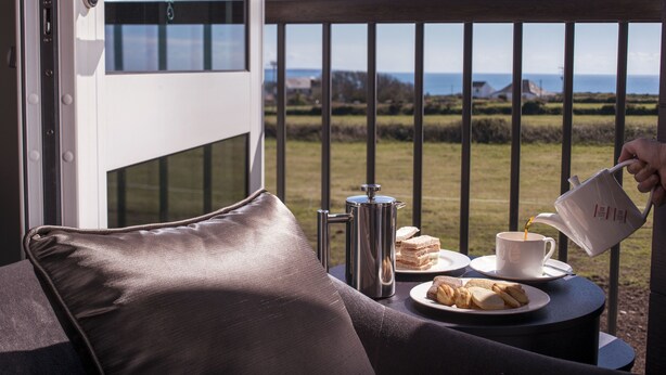 Ein Balkon mit Meerblick, eine Kaffeekanne, eine Tasse sowie ein Teller mit Sandwiches und Gebäck.