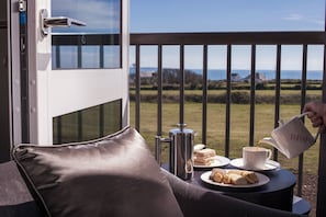 Un balcón con vista al mar, una cafetera, una taza y un plato de sándwiches y pasteles.