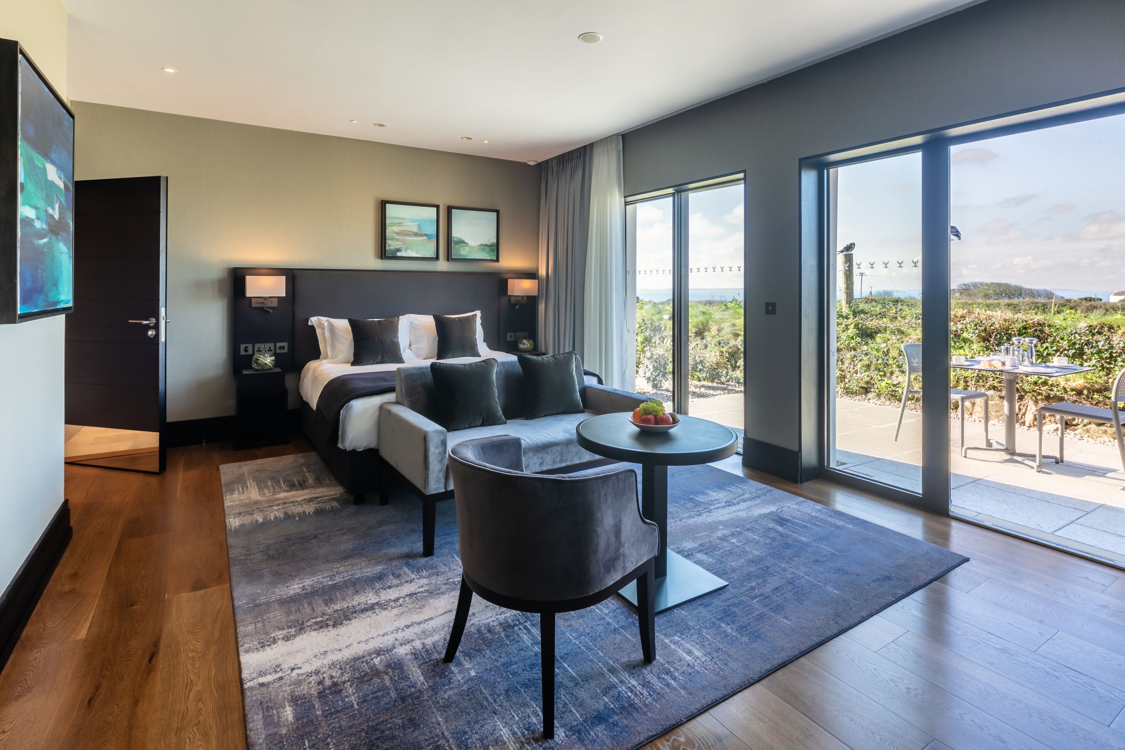 A modern hotel room with a large bed, a sofa, a small table, and a balcony with a view of the countryside.