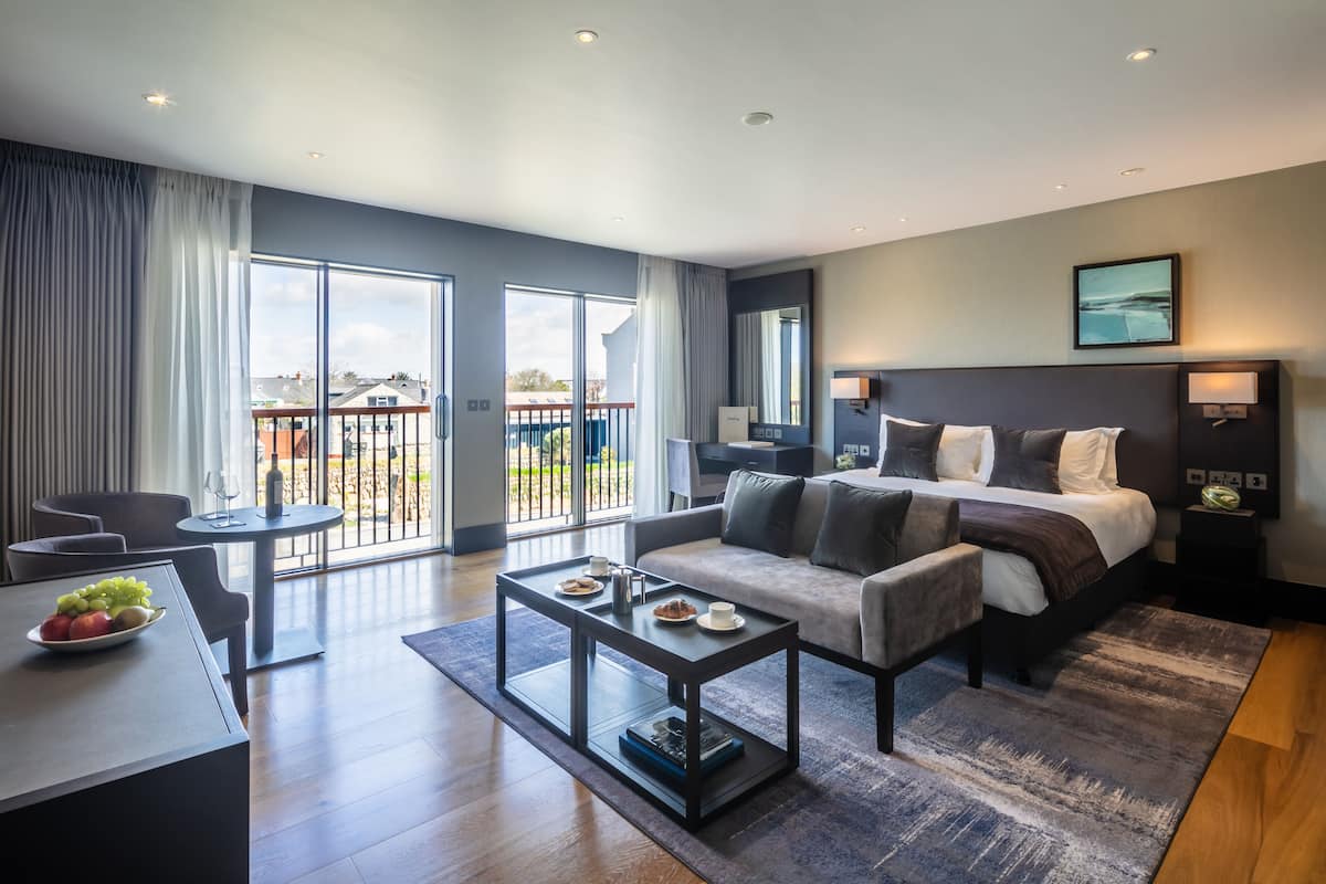 A modern hotel room with a large bed, a sofa, a dining table, and a balcony with a view.