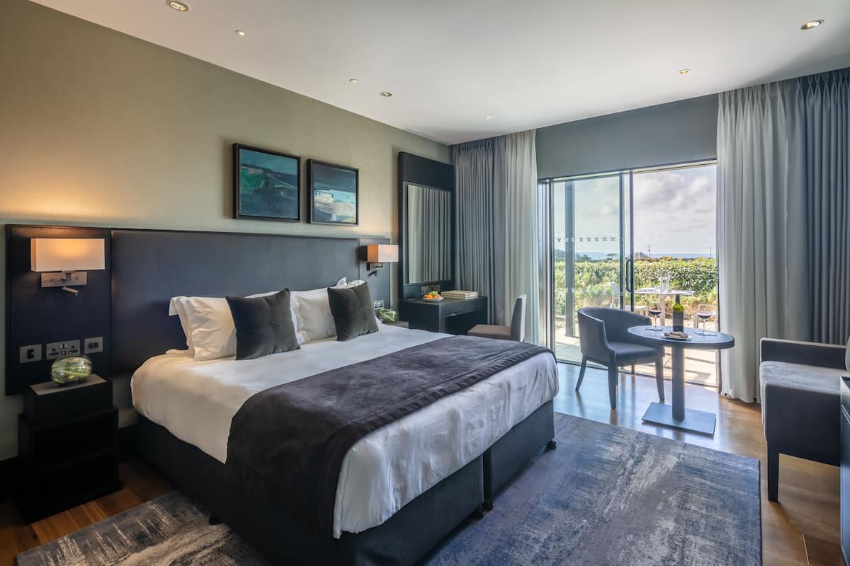 A modern hotel room with a large bed, a desk, and a balcony with a table and chairs.