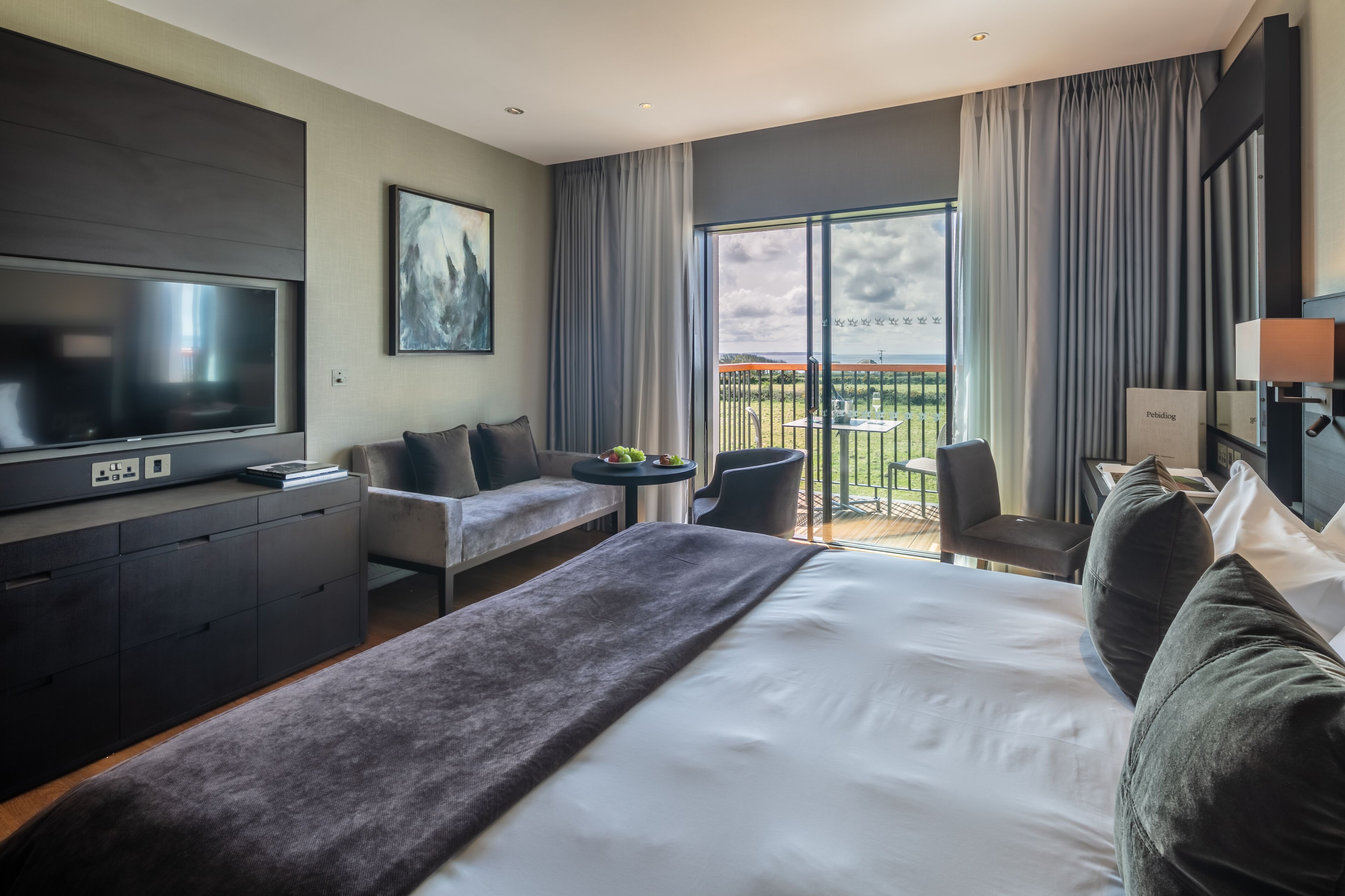 A modern hotel room with a large bed, a sofa, a TV, and a balcony with a view.