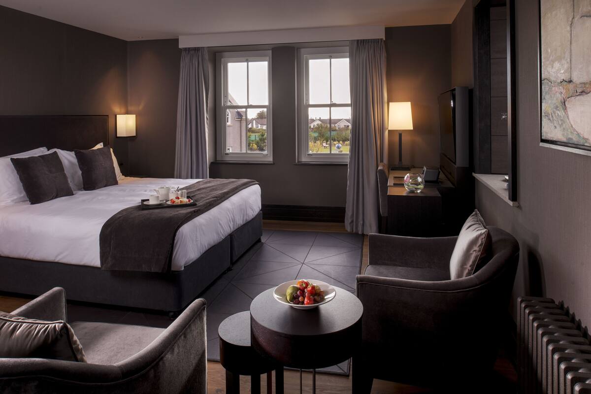 A hotel room with a large bed, two windows, a sitting area with a table, and a TV.