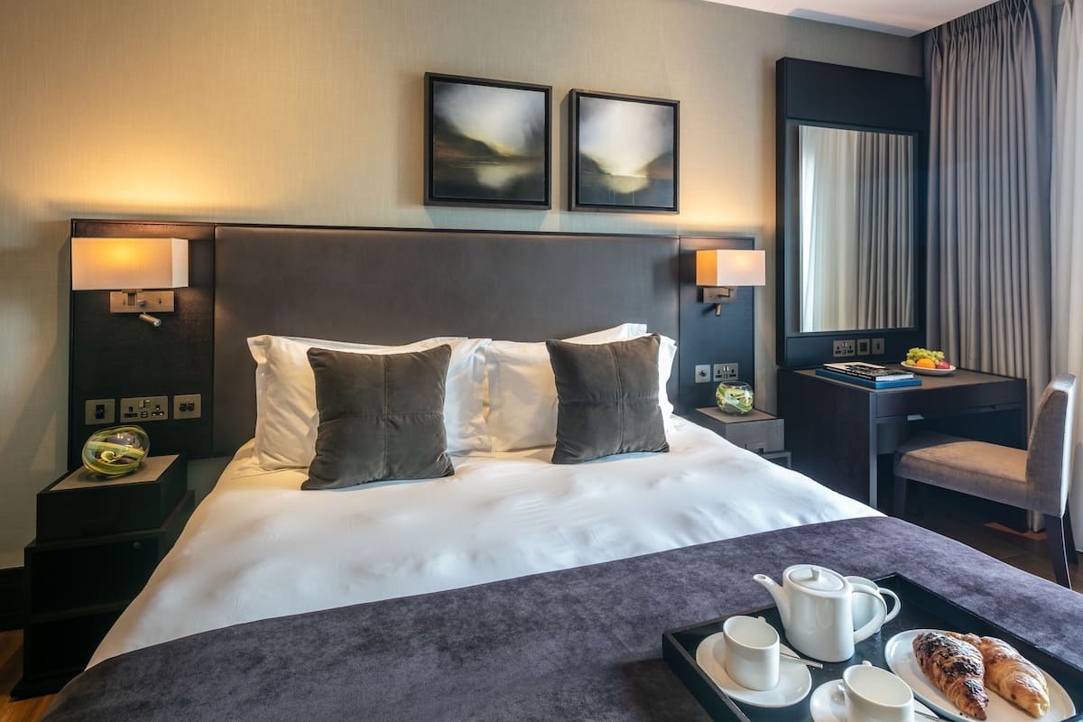 A hotel room with a large bed, two bedside lamps, a desk with a chair, and a tray with breakfast items.
