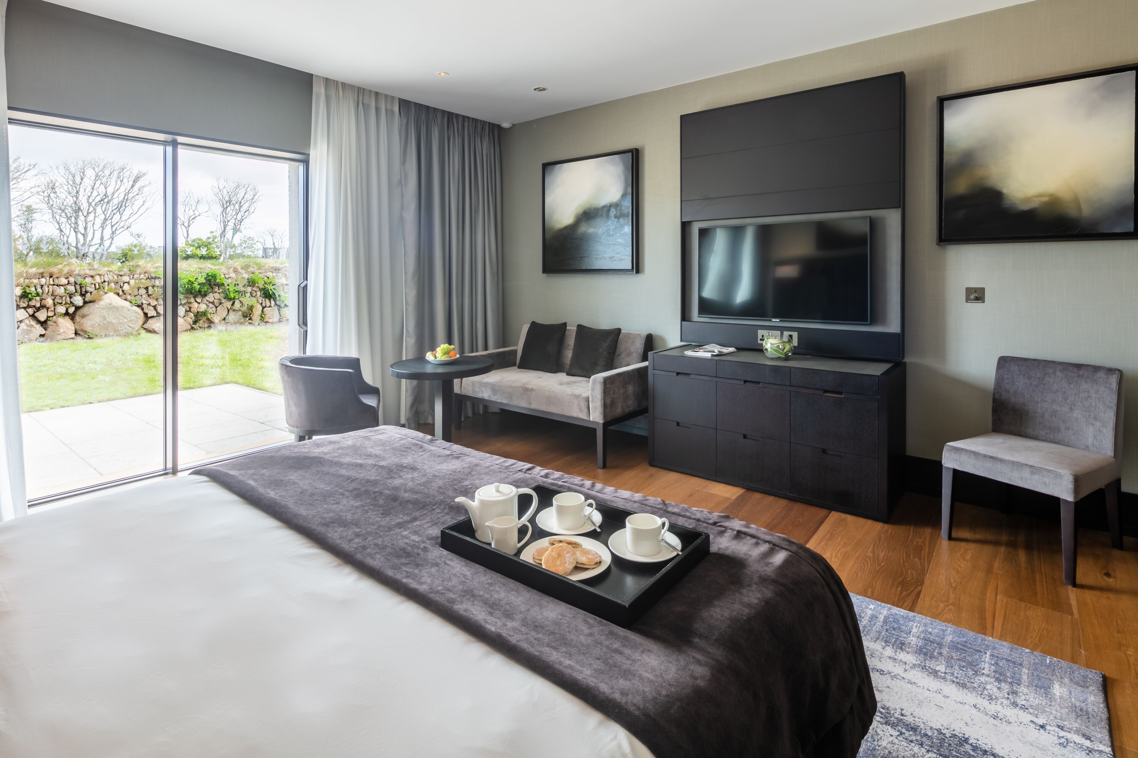 A modern hotel room with a large bed, a flat-screen TV, a seating area with a sofa and armchairs, and a dining table set with tea and pastries.