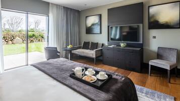A modern hotel room with a large bed, a flat-screen TV, a seating area with a sofa and armchairs, and a dining table set with tea and pastries.