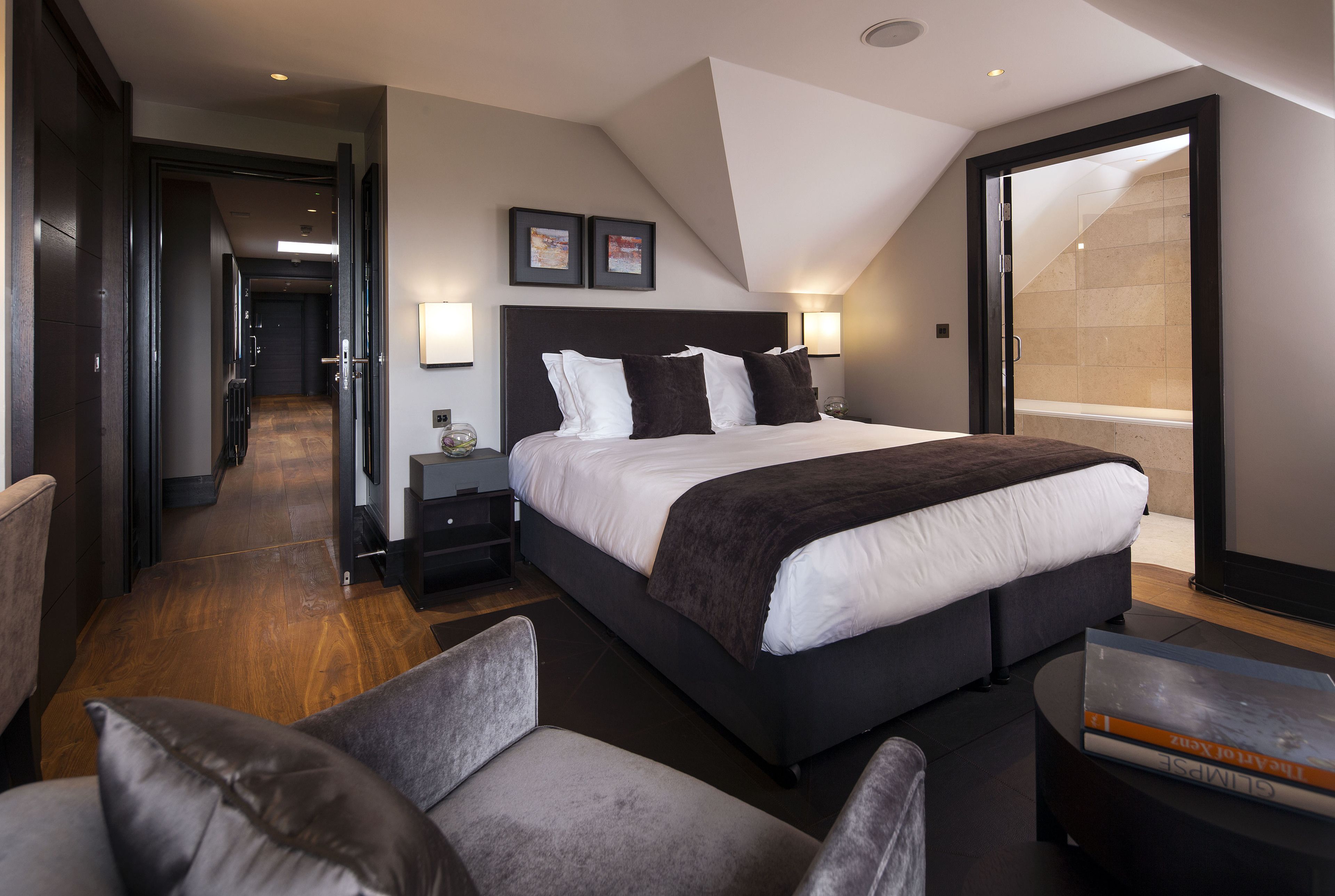 A modern hotel room with a large bed, a seating area with chairs, a bedside table, and a view of a shower area.