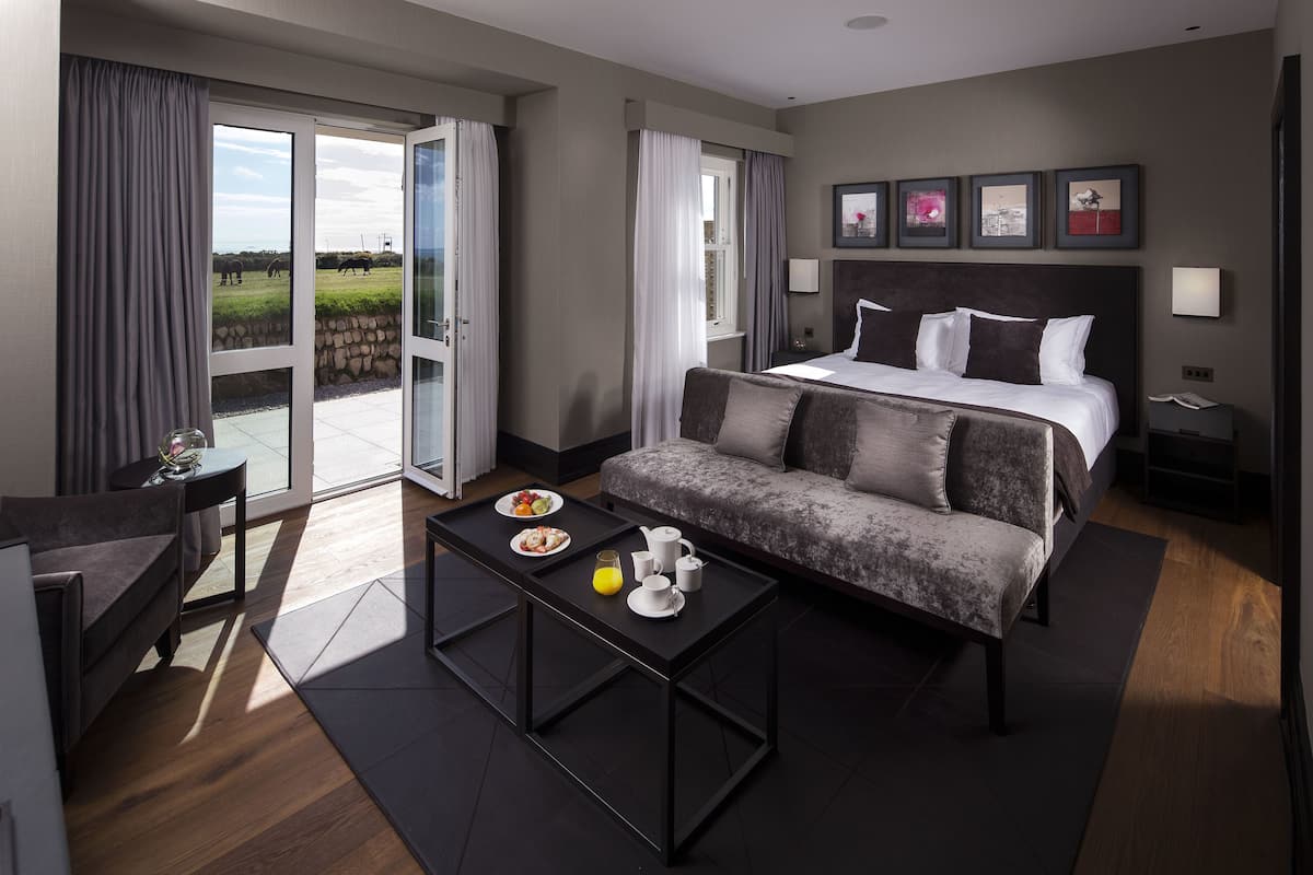 A modern hotel room with a large bed, a sofa, a dining table with breakfast, and a view of a green field through the sliding glass door.