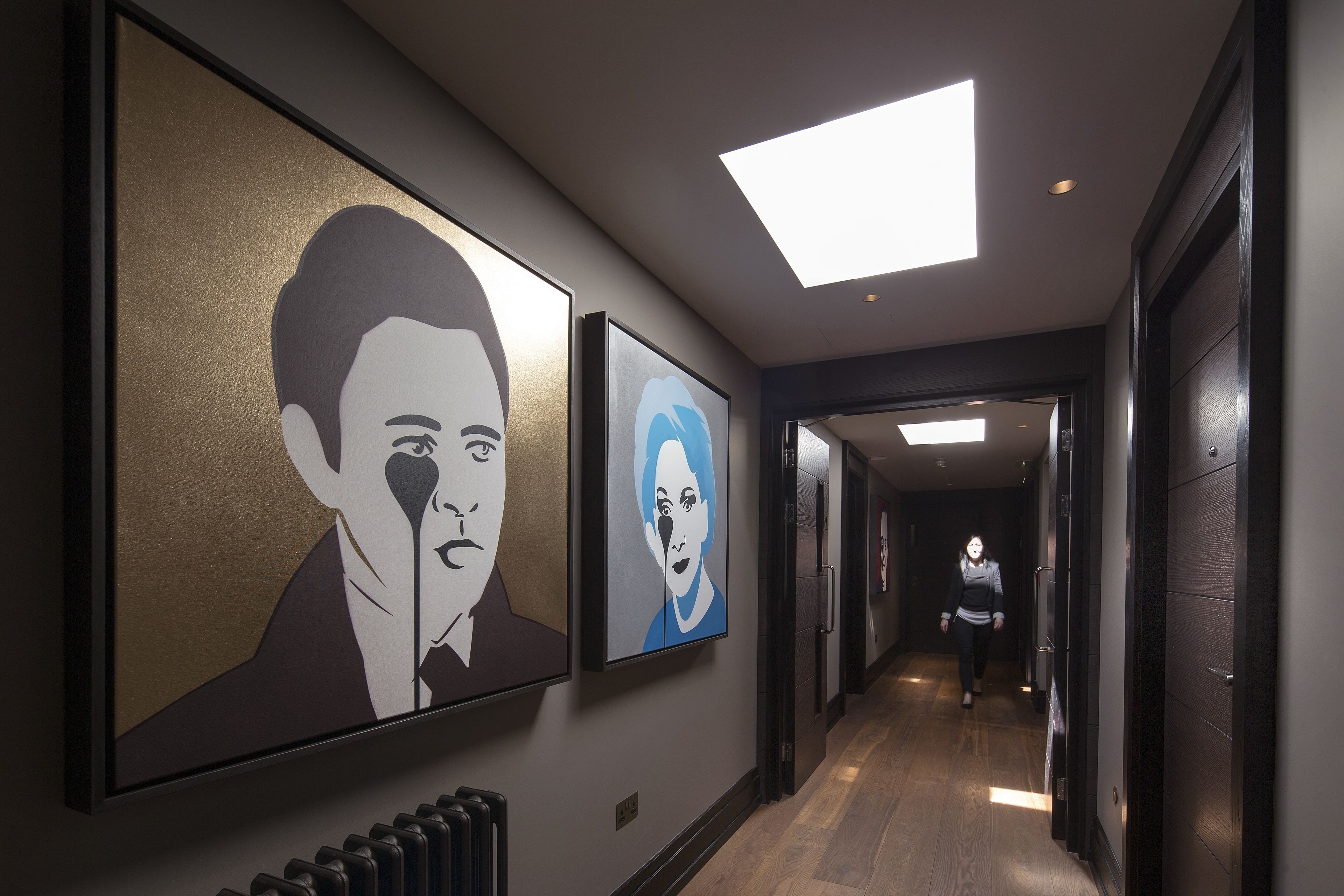 A hallway with large framed artworks on the walls, a ceiling light, and a person walking away.