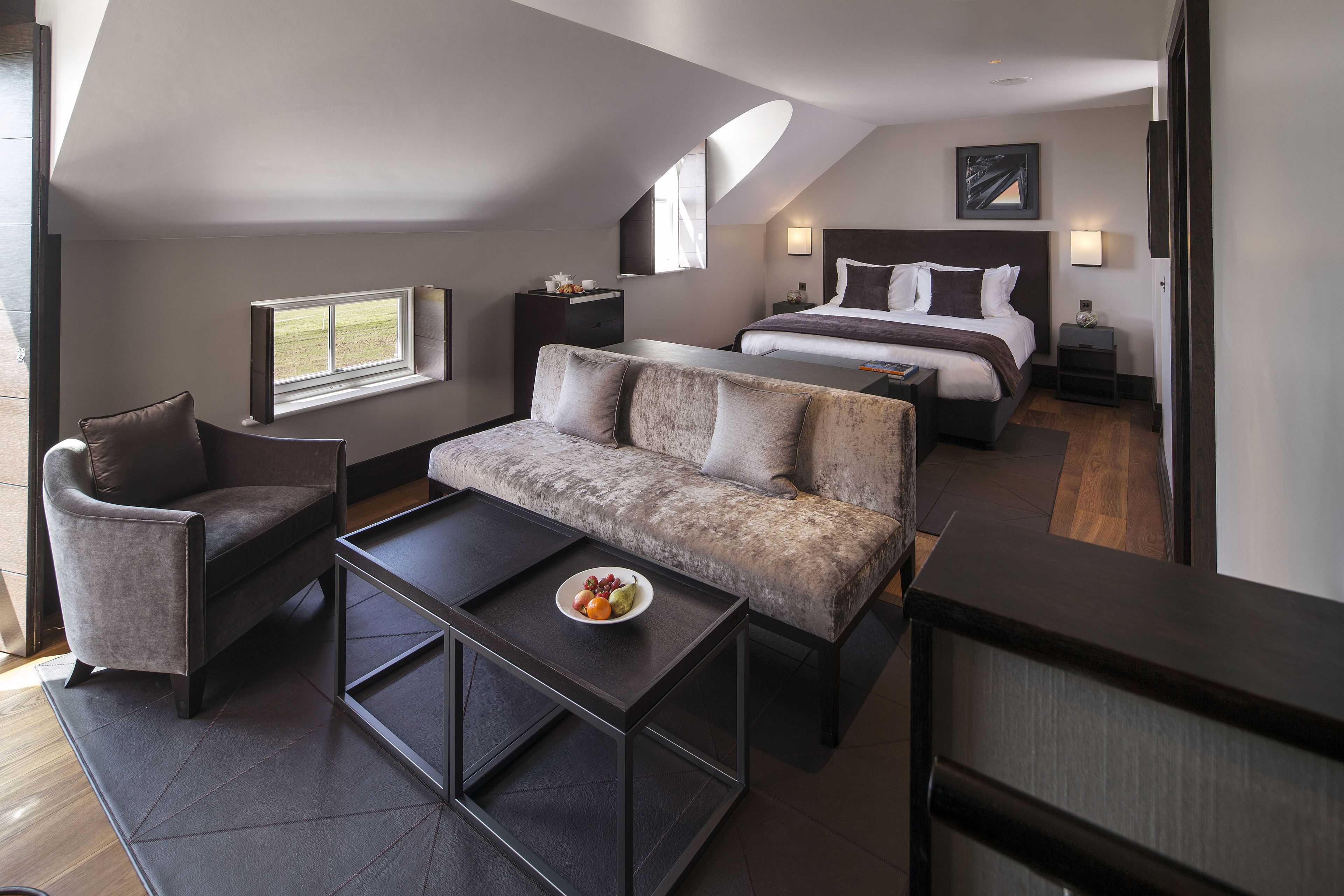 Une chambre d’hôtel moderne avec un canapé, un fauteuil et une petite table sur laquelle se trouve des fruits. On y trouve également un lit avec une tête de lit et une table de chevet avec une lampe.