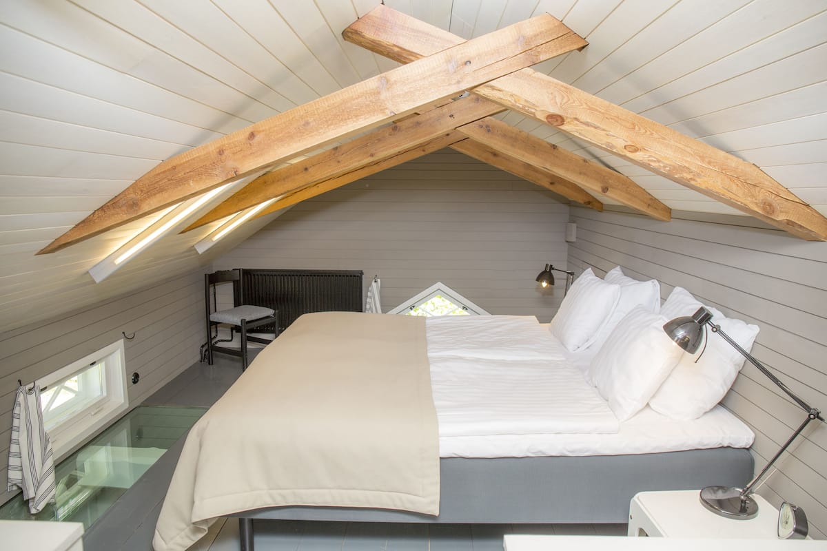 Een slaapkamer met schuine wand, een bed met wit bedlinnen, een bedlampje en een raam met zonwering.