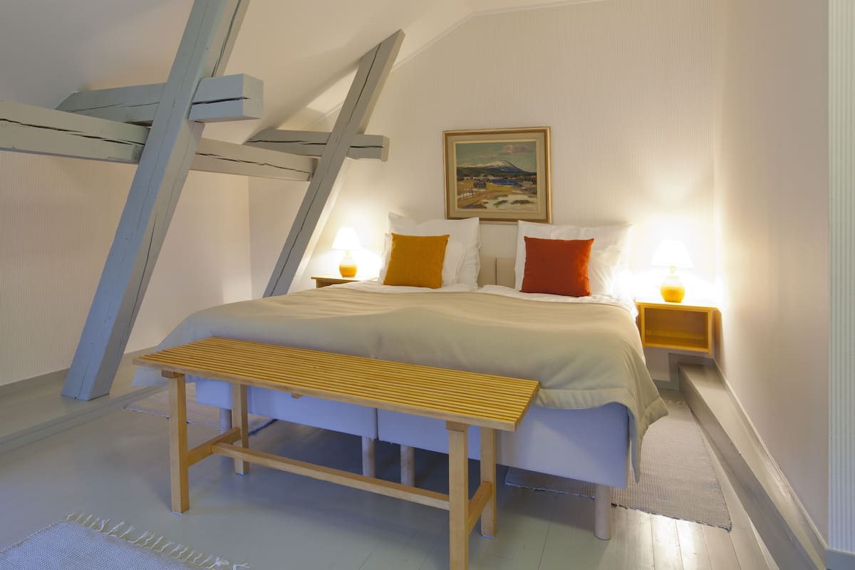 Een slaapkamer met een schuine plafond, een bed met wit en oranje beddengoed, twee bedlampen, een houten bankje en een schilderij aan de muur.