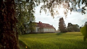 Une grande maison avec un toit rouge et des murs blancs, entourée d’un champ verdoyant et d’arbres.
