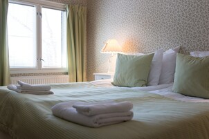 Una cama bien hecha con toallas dobladas, una lámpara de noche y una ventana con cortinas.