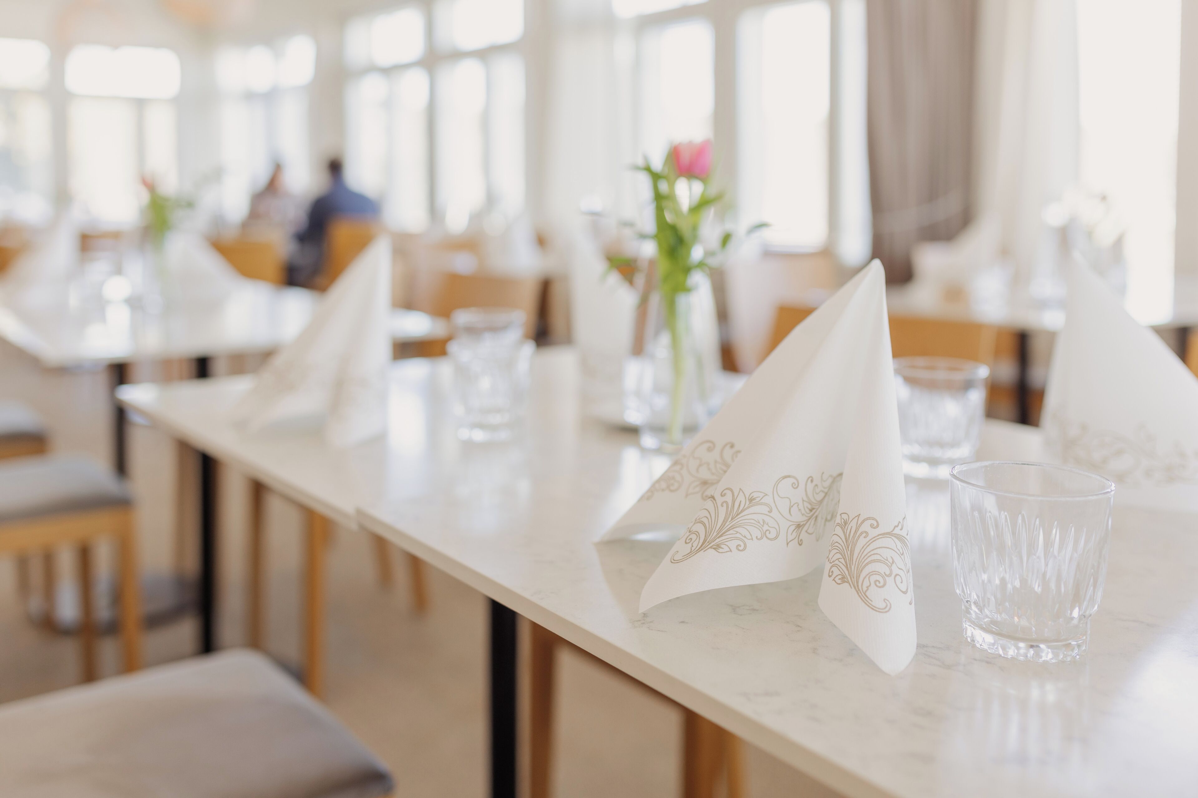 Een eetruimte met tafels, gedekt met witte servetten en heldere glazen, een vaas met bloemen en een raam op de achtergrond.