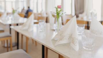 Een eetruimte met tafels, gedekt met witte servetten en heldere glazen, een vaas met bloemen en een raam op de achtergrond.