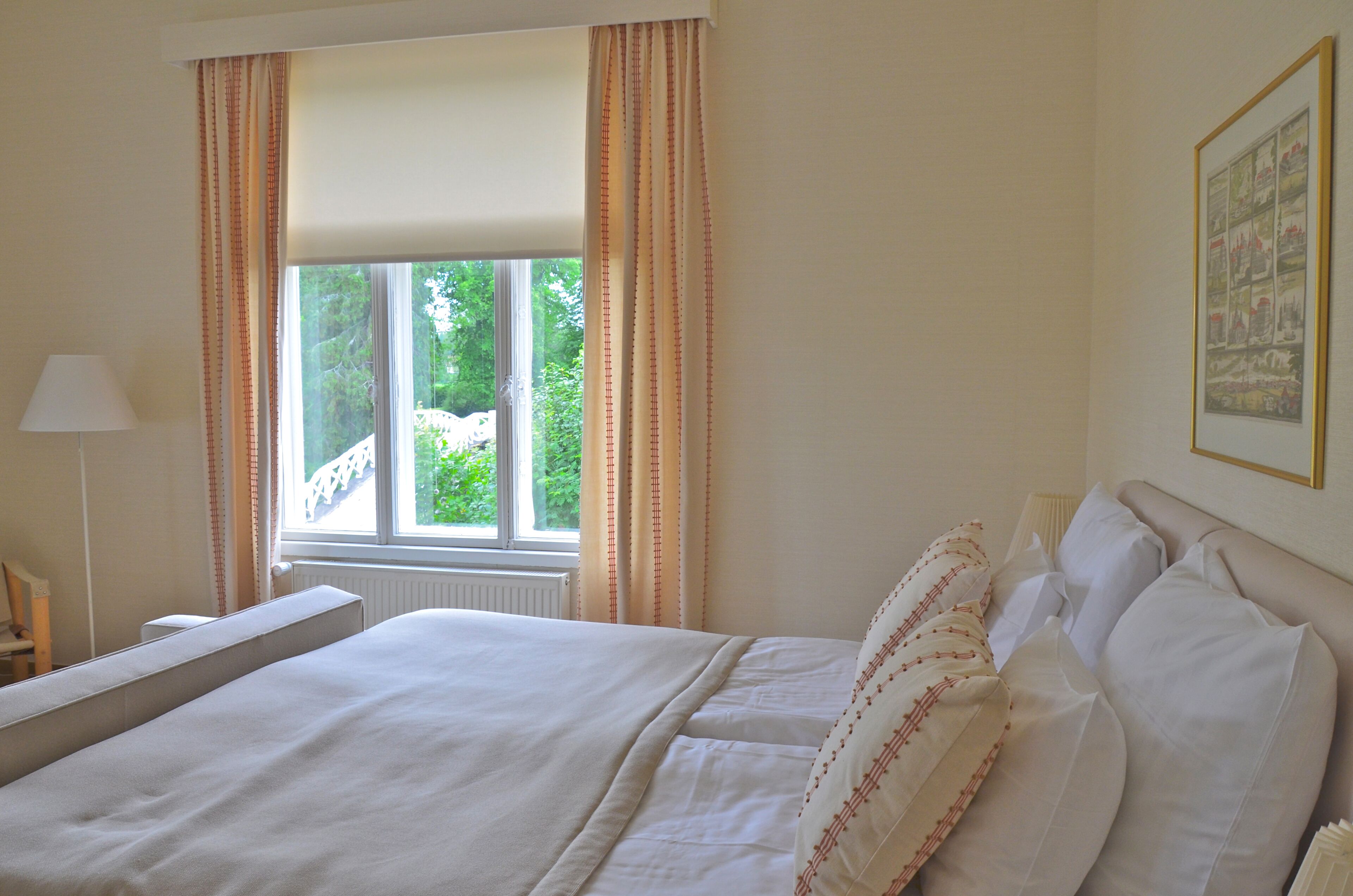 Une chambre d’hôtel avec un grand lit, une fenêtre avec des rideaux, une table de chevet avec une lampe et un tableau encadré au mur.