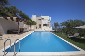 A modern pool area with a clear blue pool, a pergola, and a building with a stone facade in the background. - Villa Veghera (Kissamos)