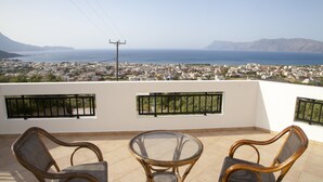 Un balcone con due poltrone in vimini e un tavolino in vetro, affacciato su una cittadina costiera con montagne sullo sfondo.