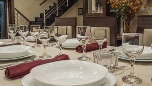 Una tavola imbandita con piatti bianchi, bicchieri trasparenti e posate, con un vaso di fiori e una scala sullo sfondo.