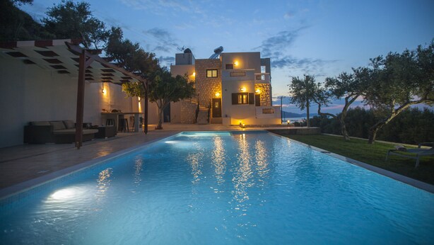 Un'area piscina moderna con piscina trasparente, zona relax coperta e un edificio con numerosi balconi e illuminazione esterna.