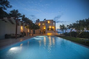 A modern poolside area with a clear pool, a covered seating area, and a building with multiple balconies and outdoor lighting. - Villa Veghera (Kissamos)