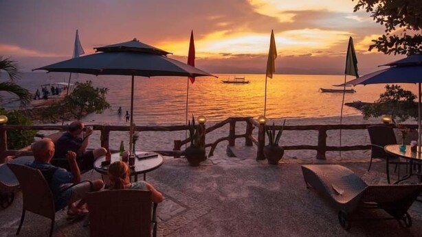 An outdoor seating area with tables and chairs, overlooking a body of water at sunset.