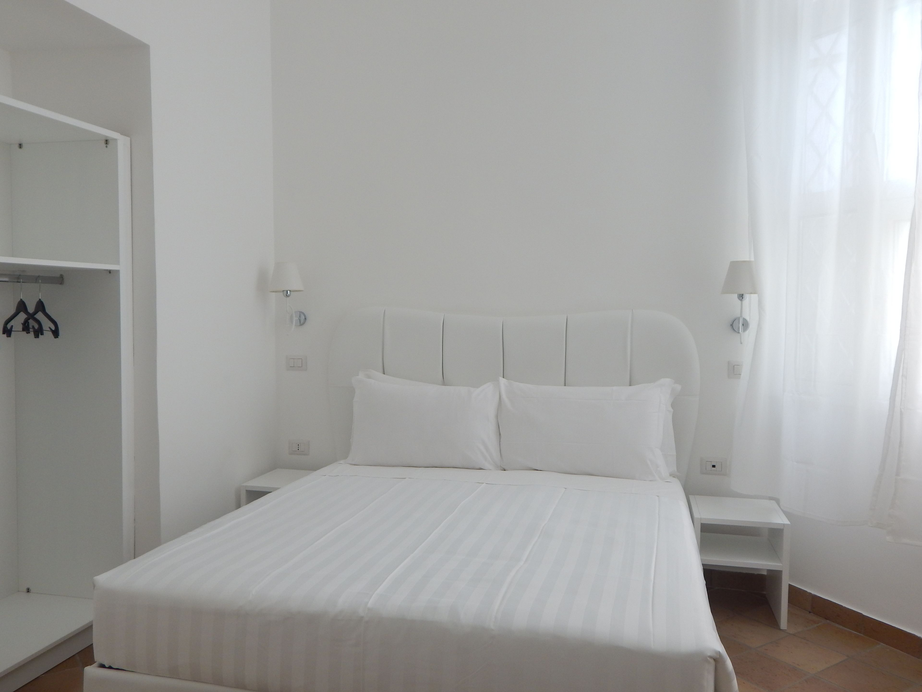 A white bedroom with a bed, two bedside tables, a wardrobe, and a window with sheer curtains.