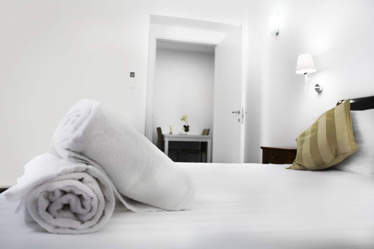 Ein Hotelzimmer mit einem ordentlich bezogenem Bett, einem aufgerollten weißen Handtuch, einem kleinen Tisch mit einer Vase und einer Wandleuchte.