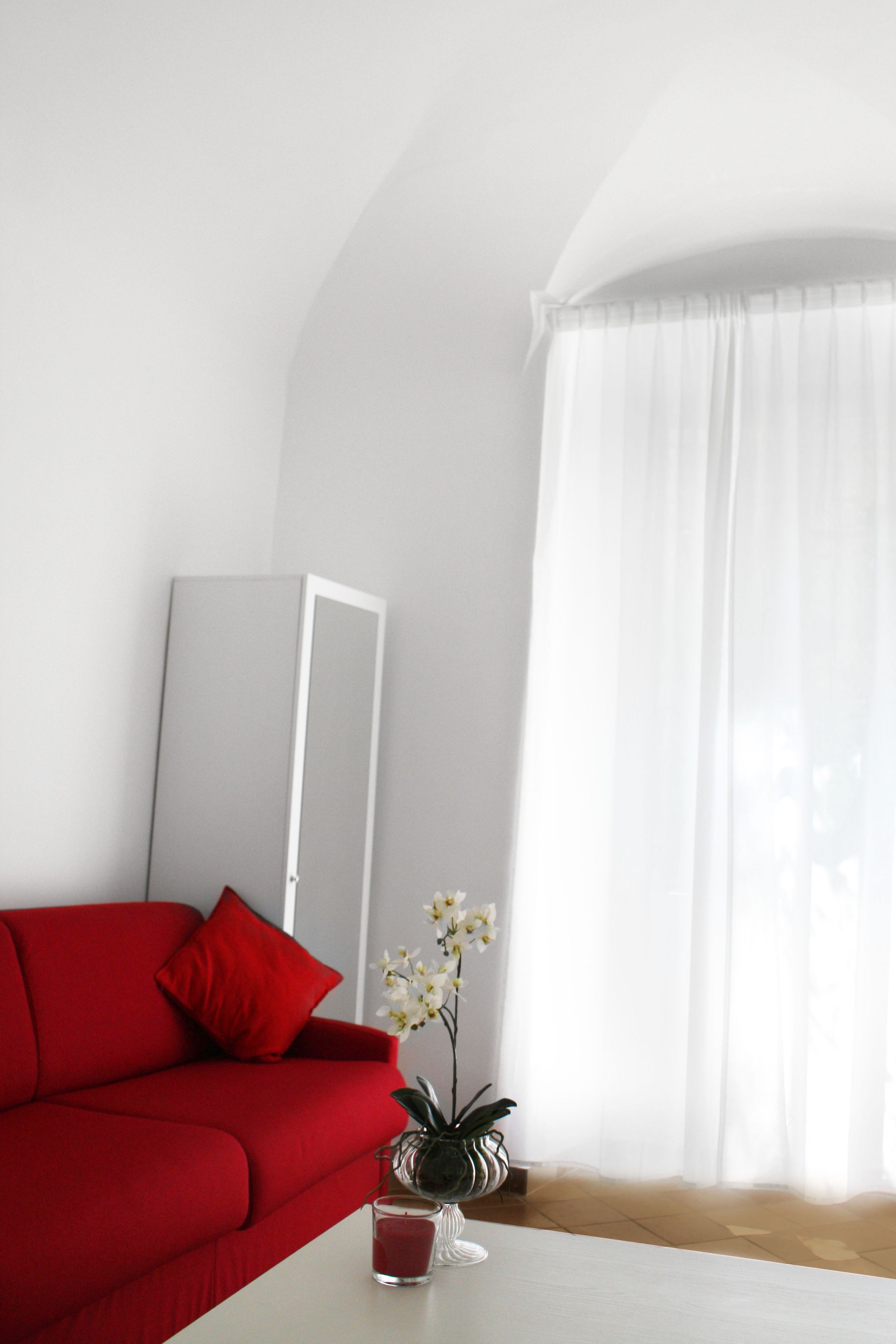 A room with a red sofa, a white table, a potted plant, and a white curtain.
