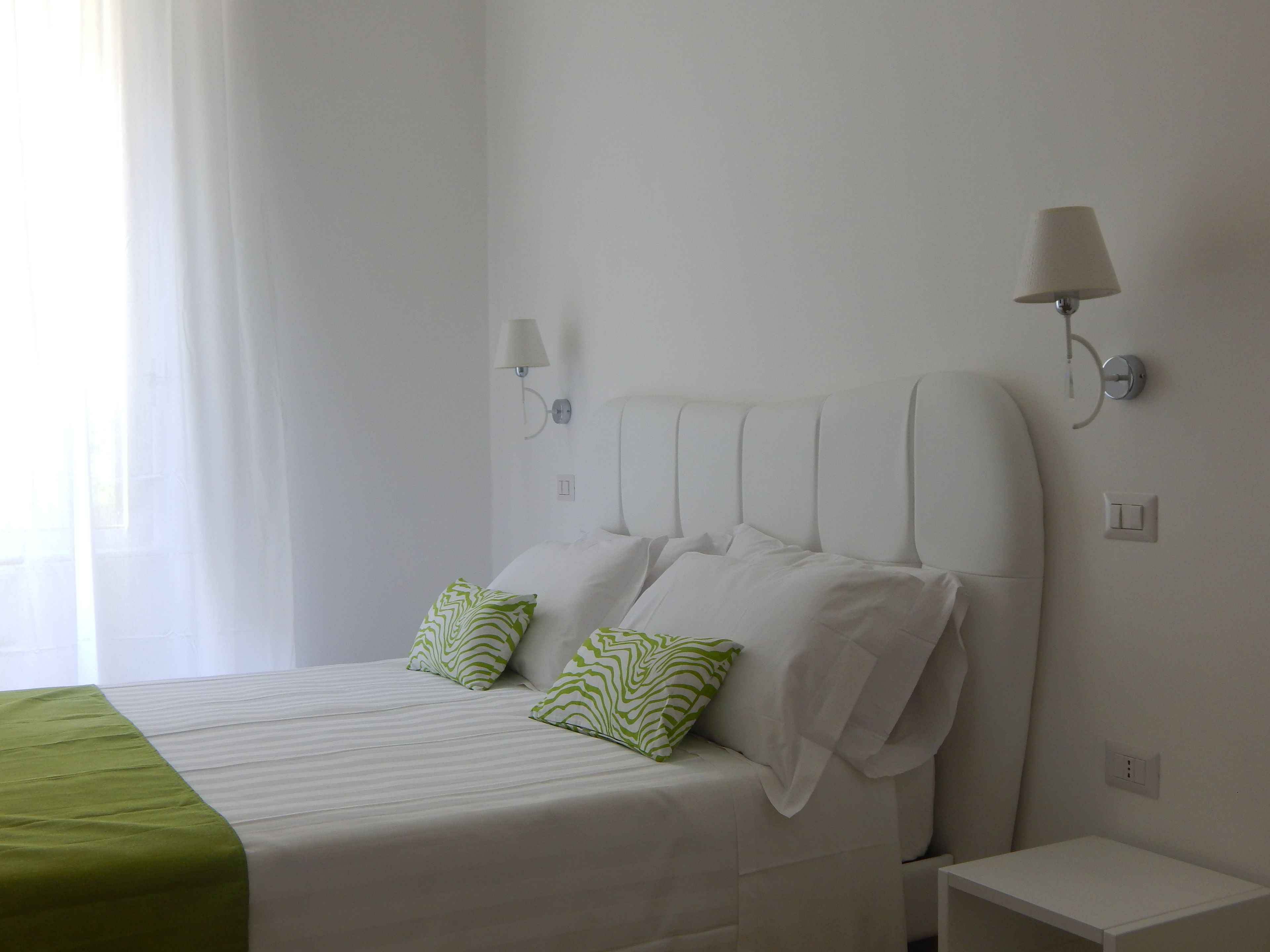 Cama bem arrumada com roupa de cama branca e duas almofadas verdes estampadas, mesa de cabeceira e abajur fixado na parede.