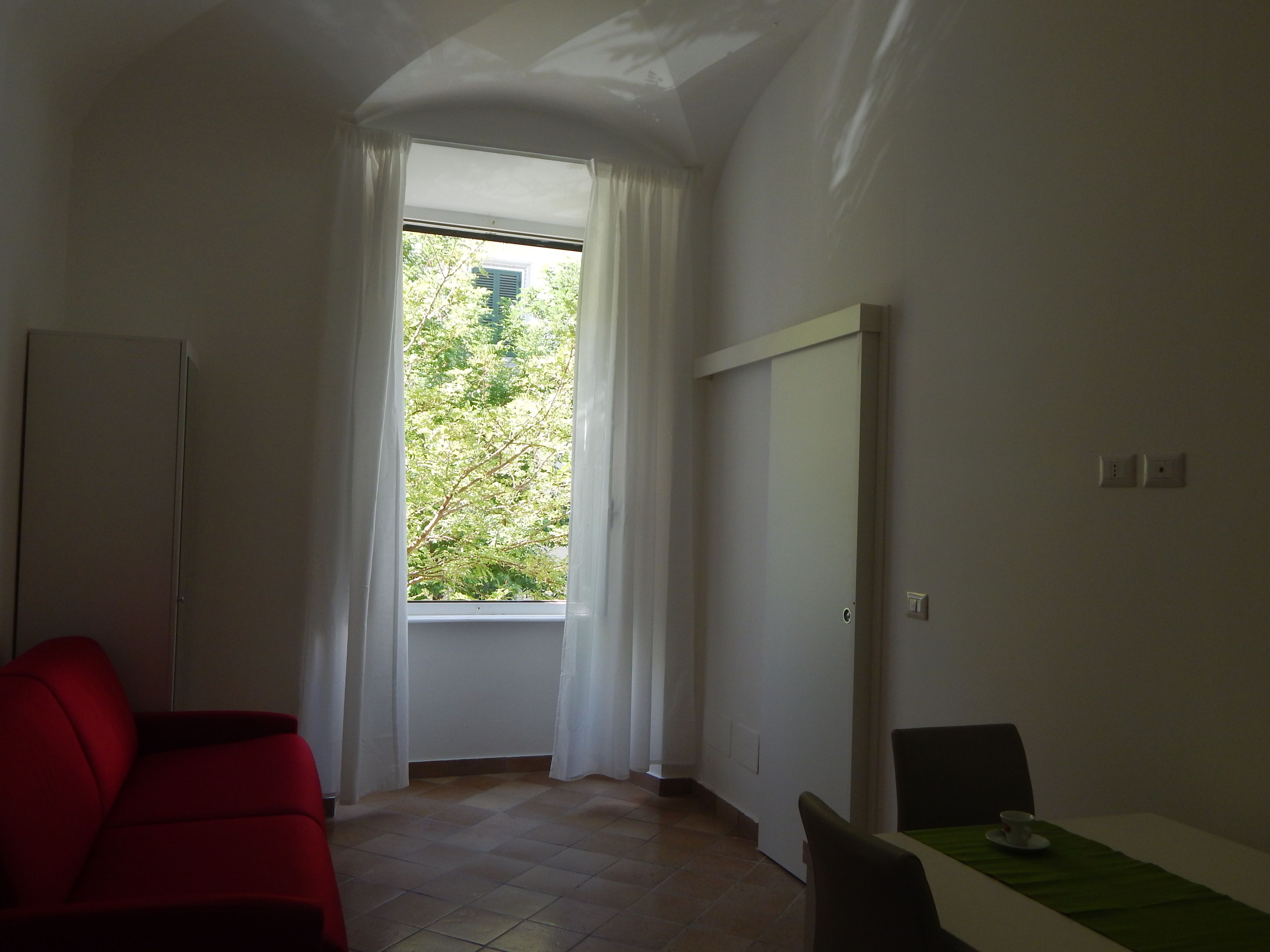 A room with a large window, a red sofa, a small table with a cup, and a white cabinet.