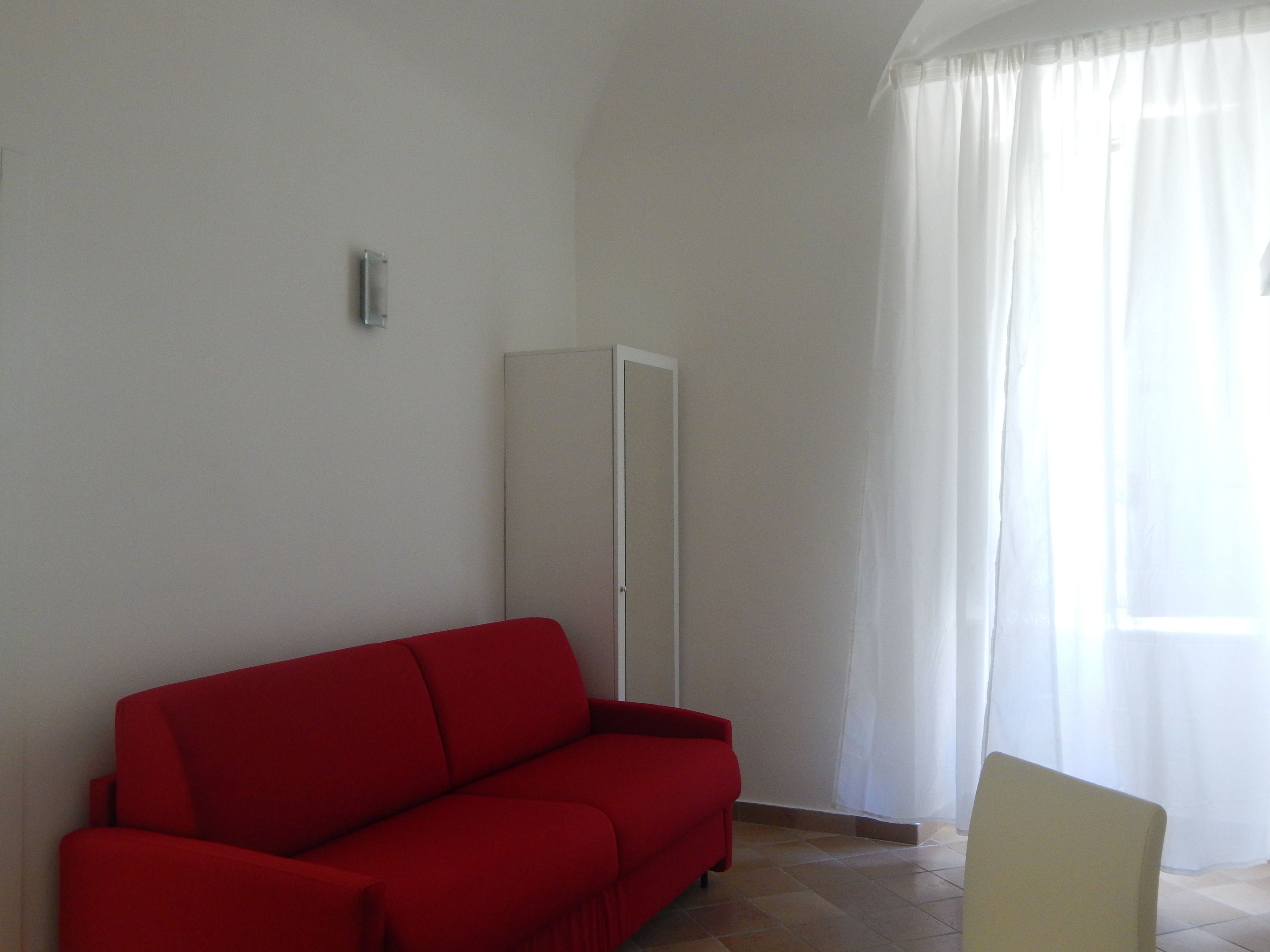A room with a red sofa, a white wardrobe, and a window with sheer curtains.