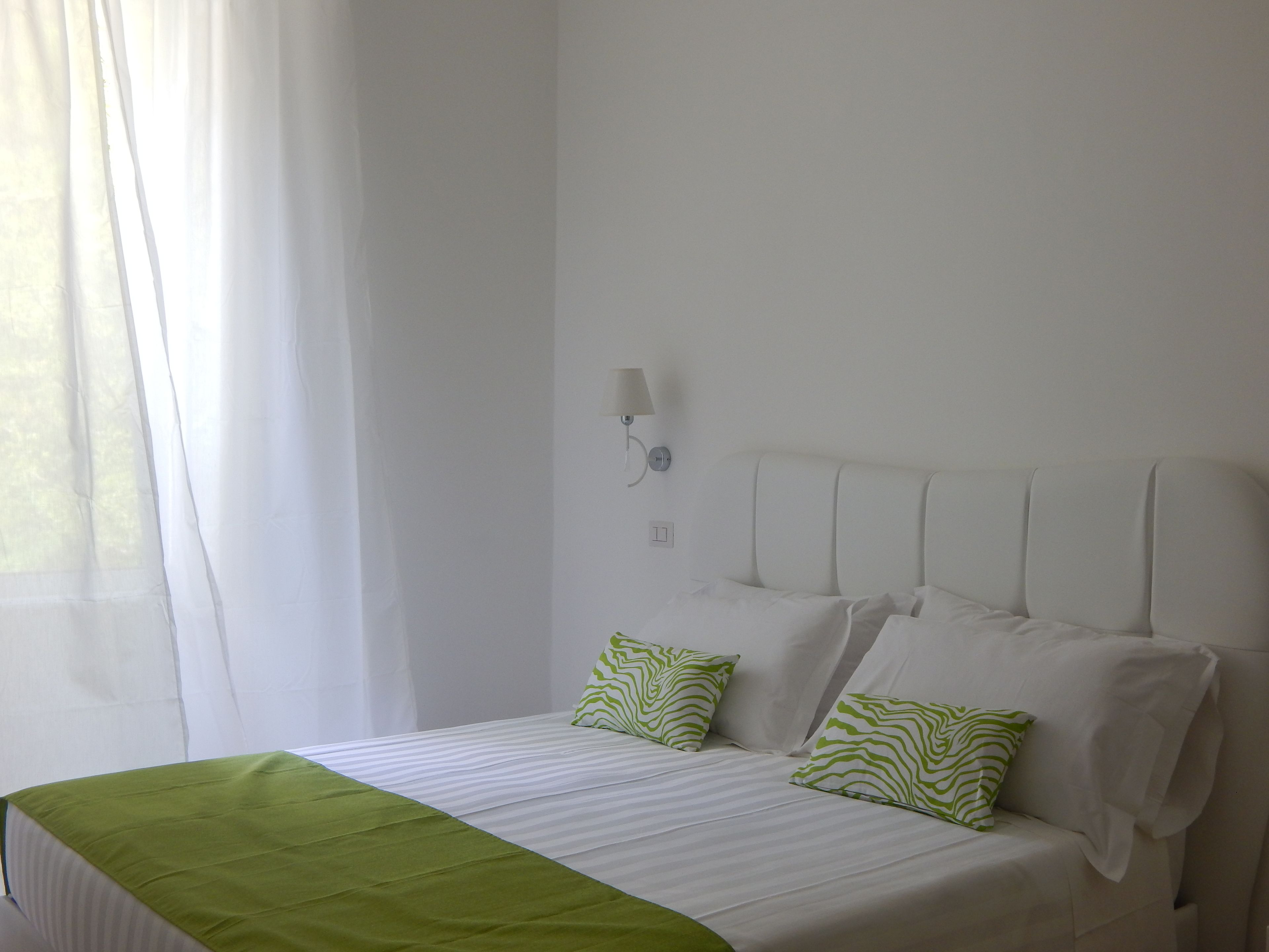 A hotel room with a bed, a green throw blanket, white curtains, and a wall-mounted lamp.