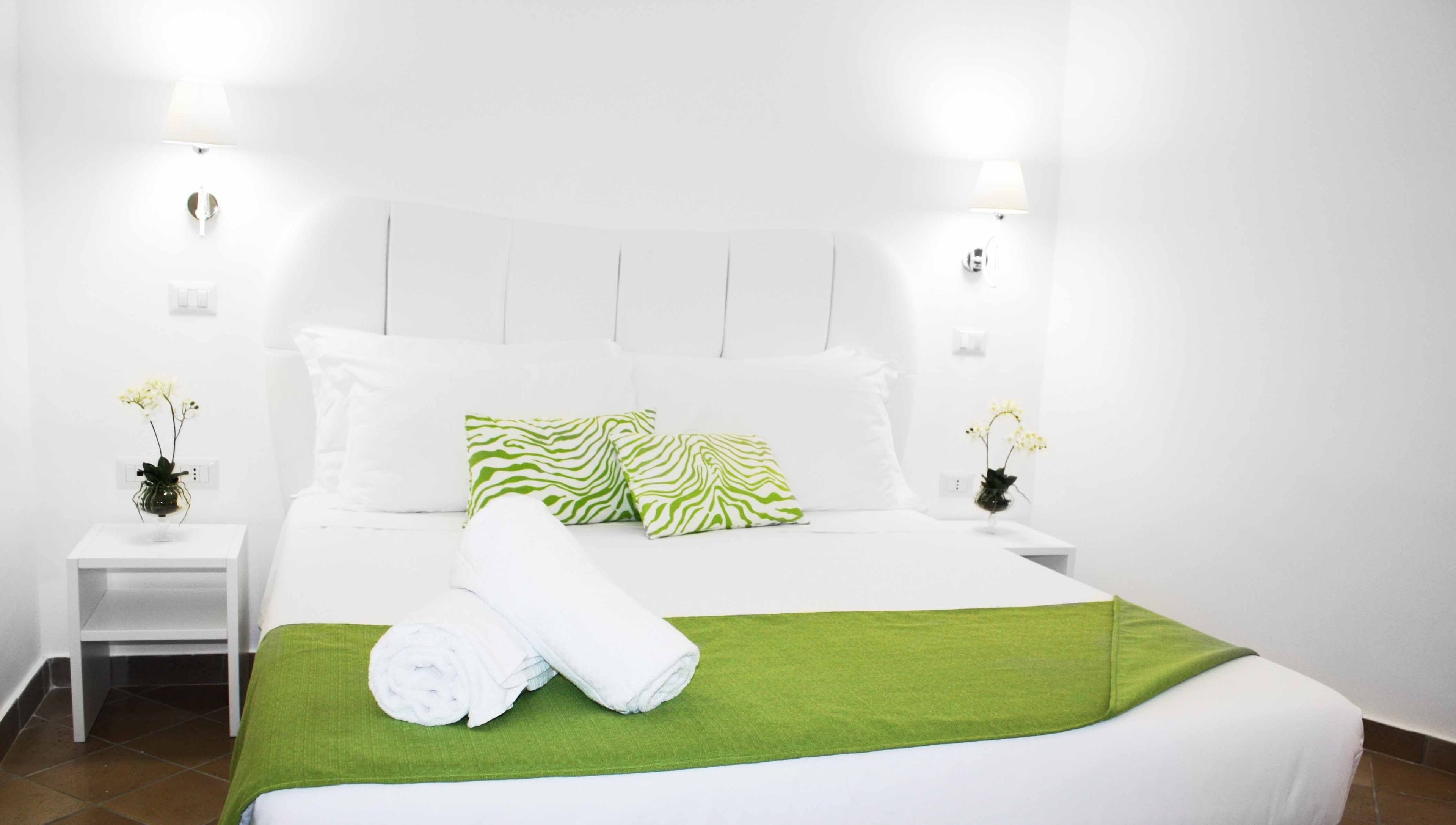 A neatly made bed with white linens and a green throw blanket, flanked by two white nightstands with lamps and small vases of flowers.
