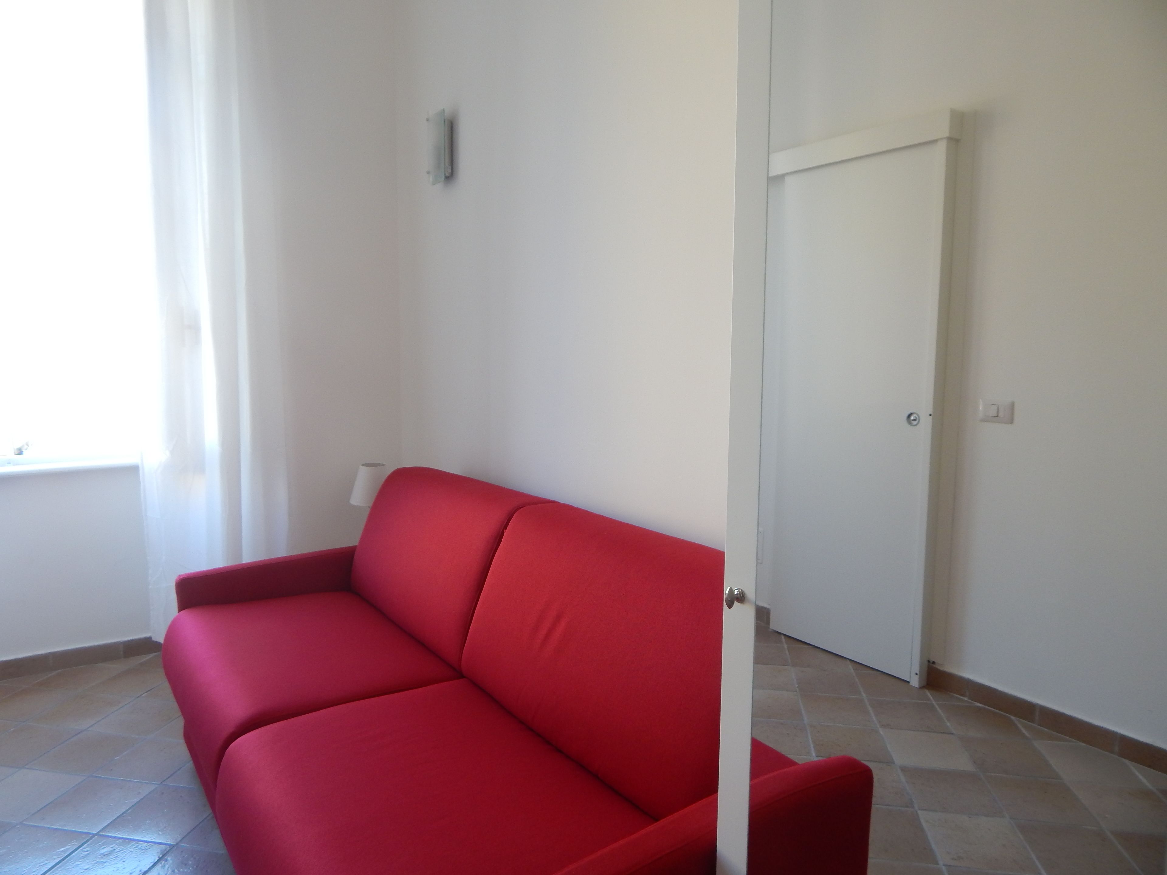 A red sofa in a room with white walls, a tiled floor, and a closed door.