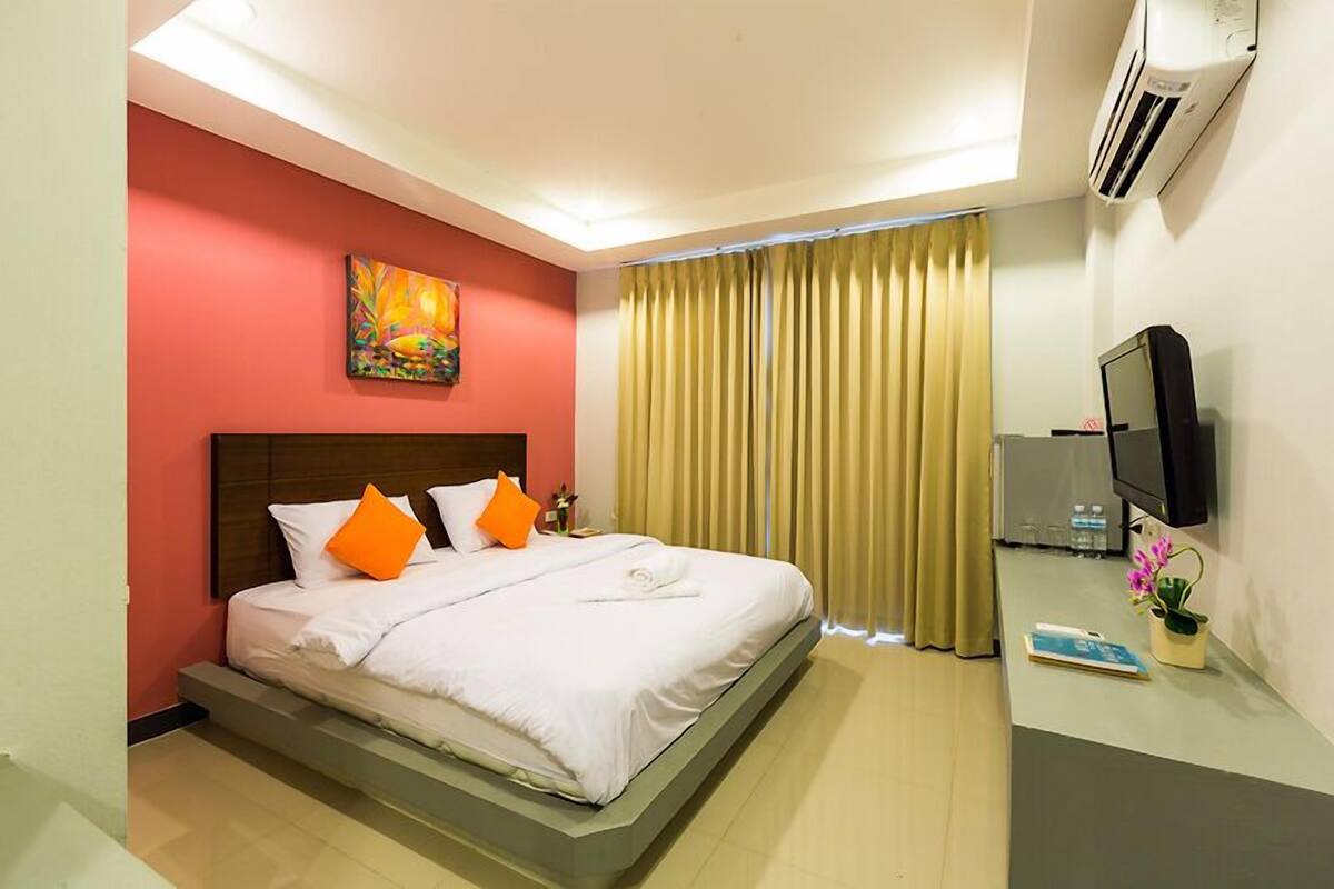 A hotel room with a bed, a flat-screen TV, an air conditioner, and a window with curtains.