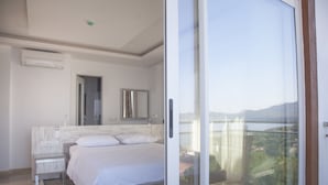 A modern bedroom with a large bed, a nightstand, a mirror, and a sliding glass door leading to a balcony with a view of the landscape. - Narr Twin (Kas)