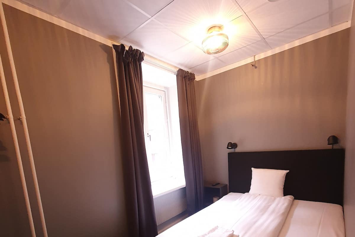 A hotel room with a neatly made bed, a bedside table, a lamp, and a window with curtains.