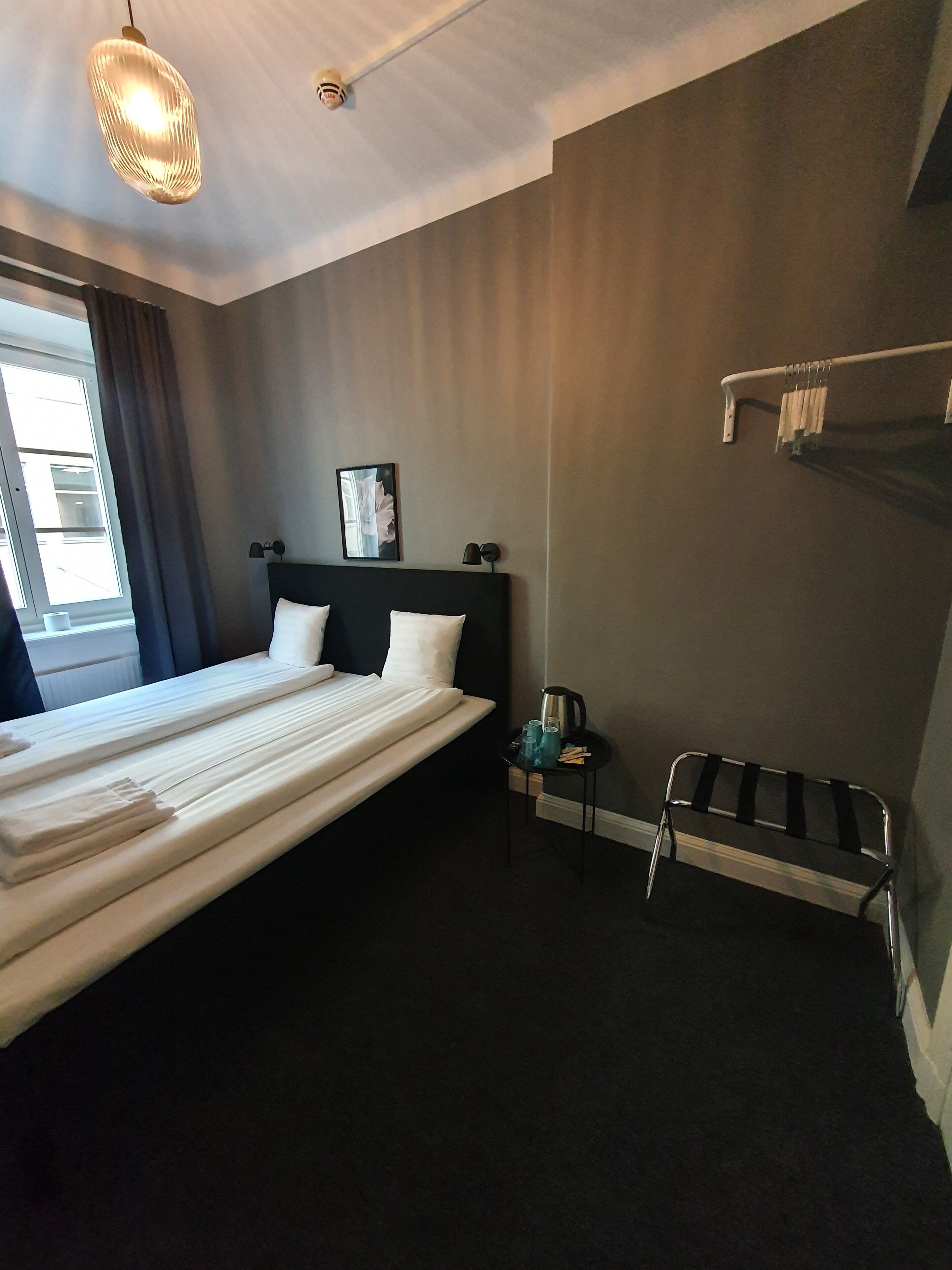 A hotel room with a large bed, a bedside table with a kettle, a chair, a coat rack, and a window with curtains.