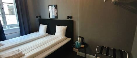 A hotel room with a large bed, a bedside table with a kettle, a chair, a coat rack, and a window with curtains.