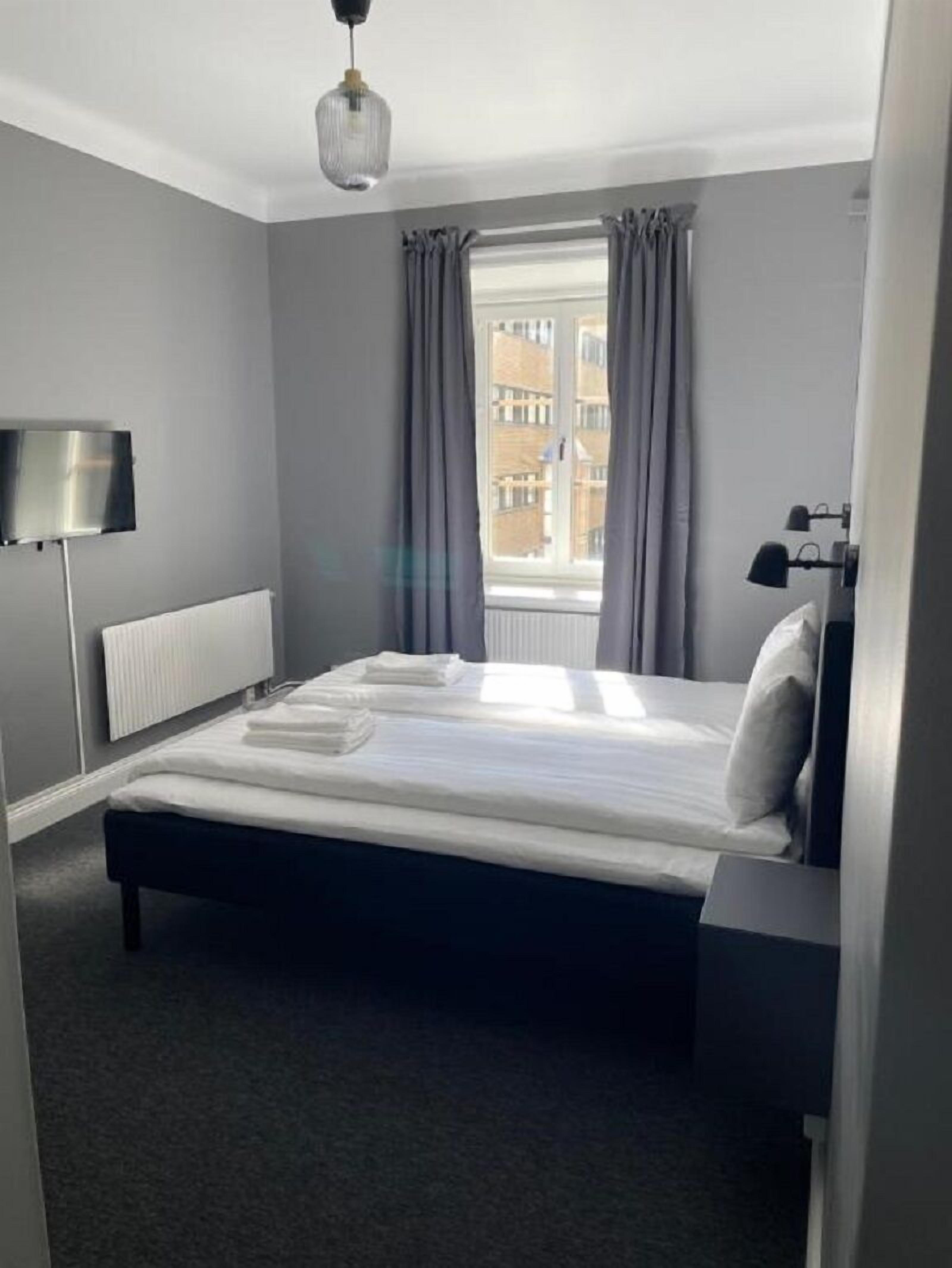 A hotel room with a bed, a bedside table, a lamp, a window with curtains, and a view of buildings outside.