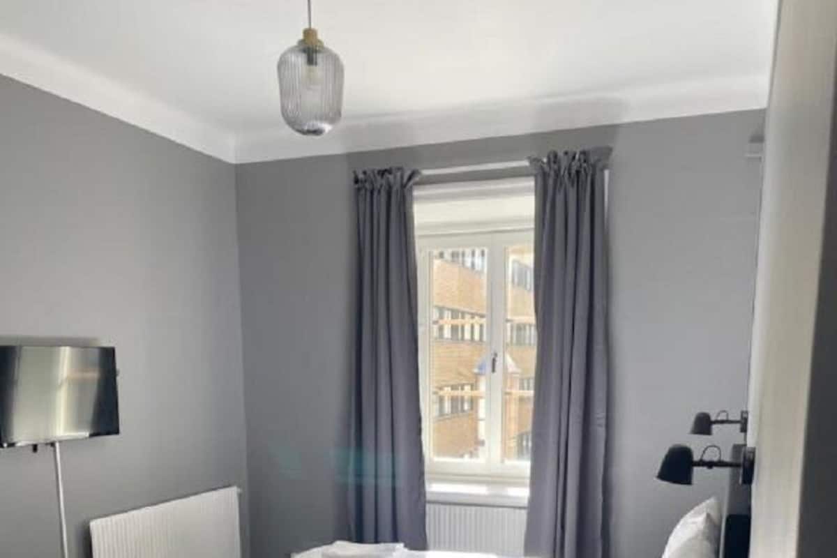 A hotel room with a bed, a bedside table, a lamp, a window with curtains, and a view of buildings outside.