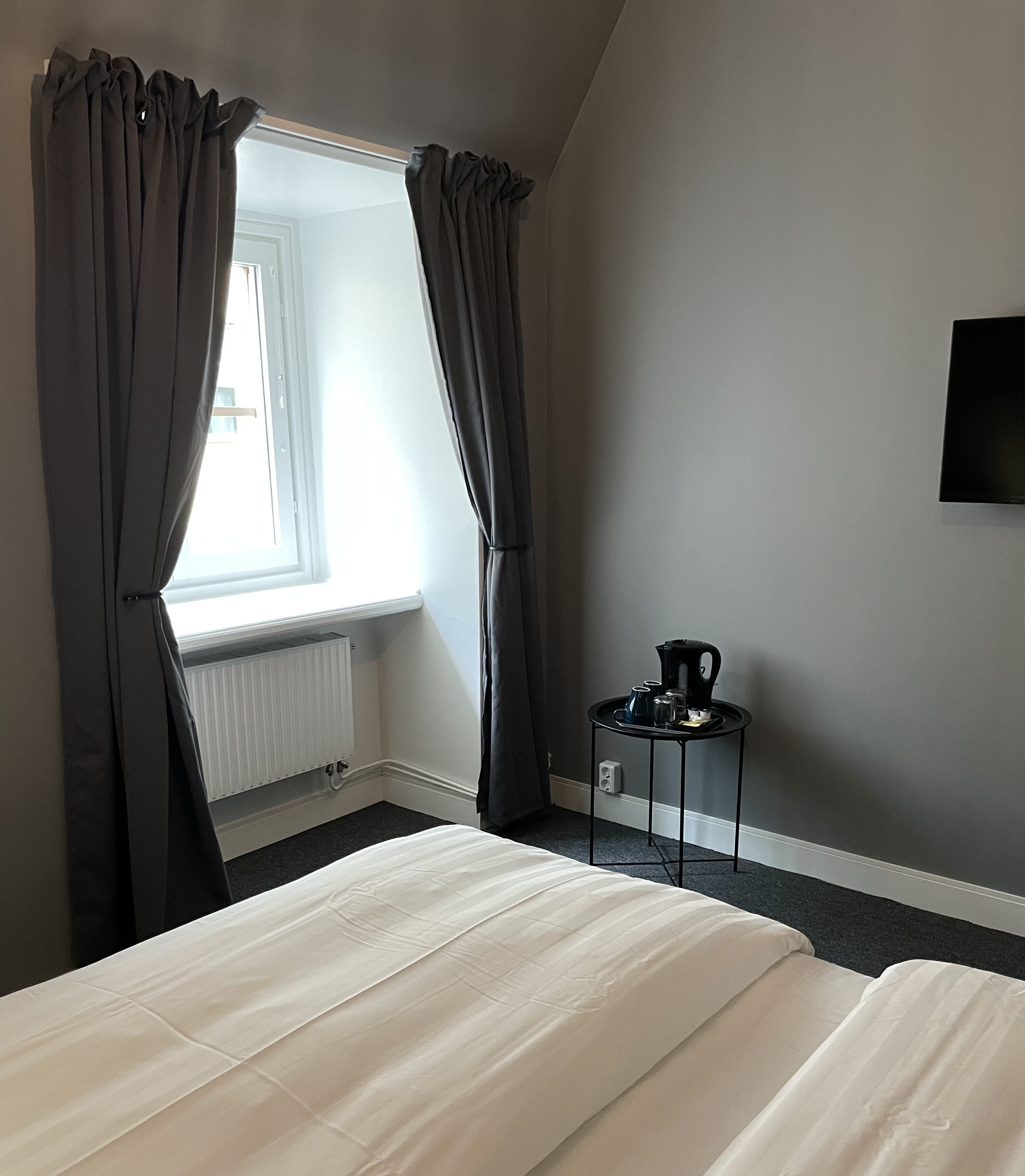 A bedroom with a bed, a window with curtains, a radiator, and a small table with a kettle.